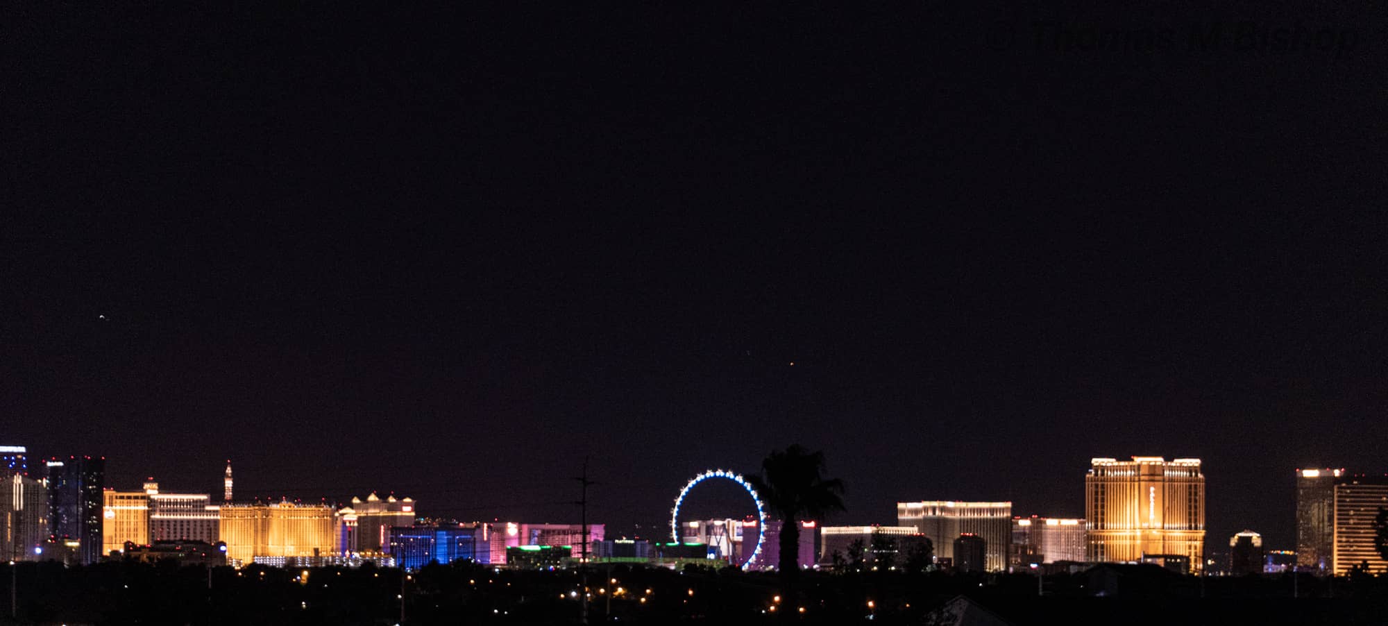 The Las Vegas Strip at night seen from a distance, a low line of lit casino towers and a large illuminated observation wheel under a dark sky