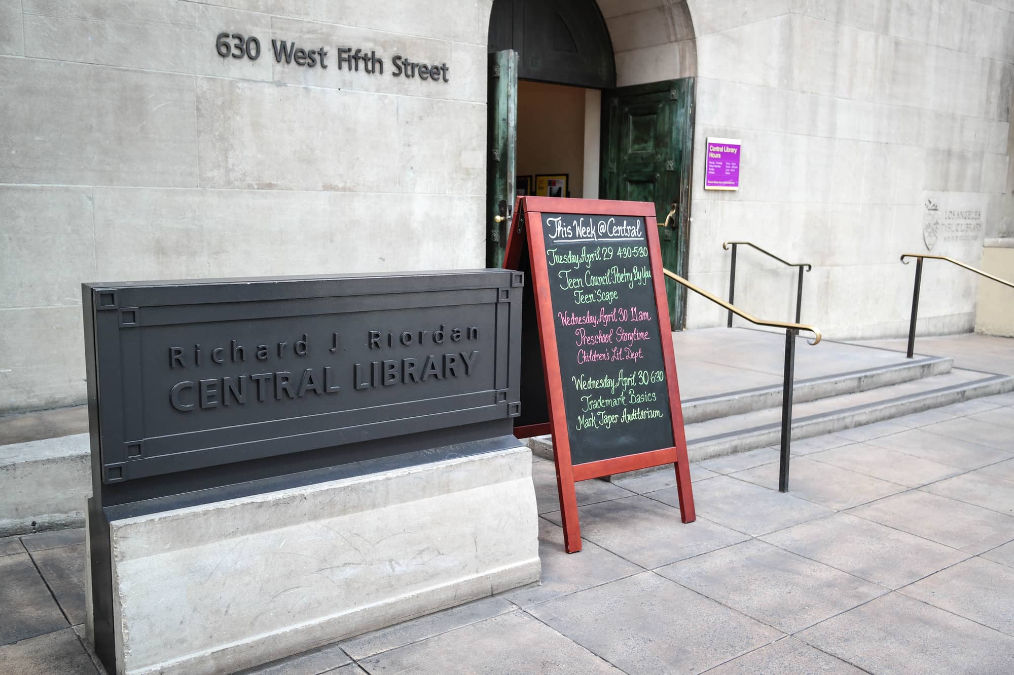 The entrance to the Richard J. Riordan Central Library at 630 West Fifth Street in Los Angeles, with a stone sign, a chalkboard of events and open green doors