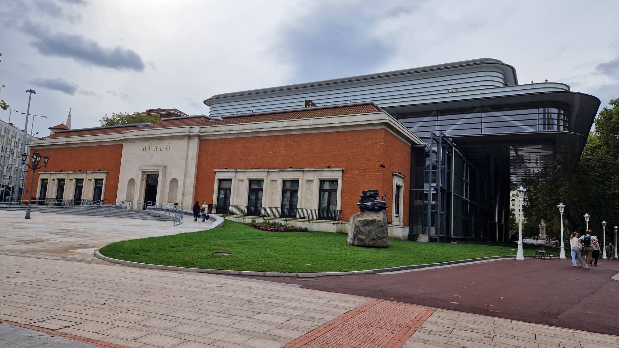 The Bilbao Fine Arts Museum extension seen from the plaza: the 1945 red-brick museum with its stone MVSEO portal, the new curved metal-clad gallery rising behind it and its glazed atrium to the right