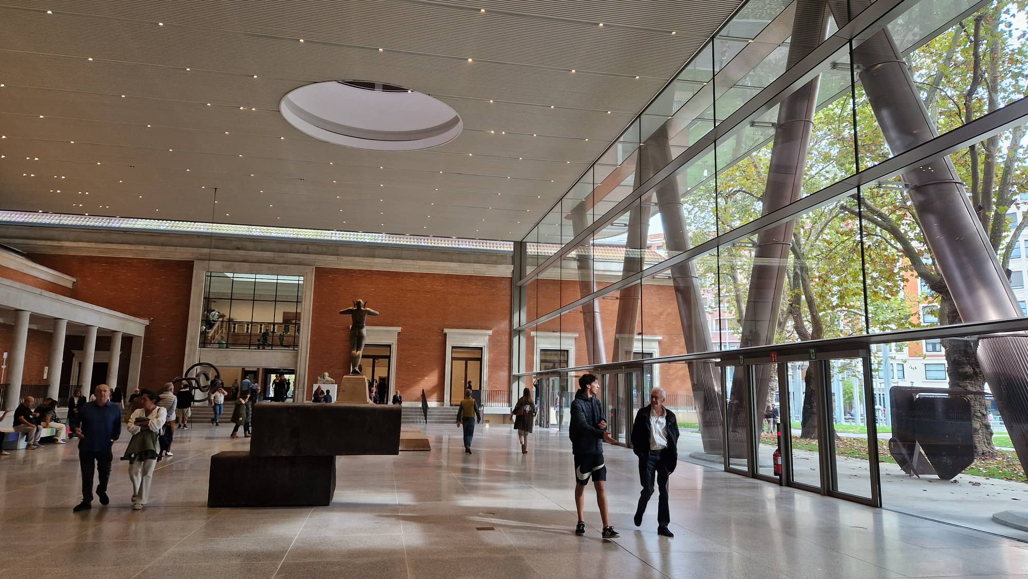The covered Arriaga atrium of the Bilbao Fine Arts Museum: a circular oculus in the ceiling, the 1945 facade as an interior wall, steel columns behind glass and visitors crossing the stone floor