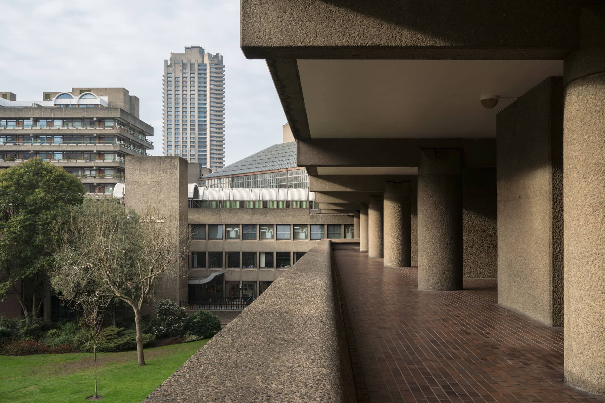 A raised concrete walkway at the Barbican Estate in London, with rough, textured concrete columns, brown tiles and a residential tower beyond