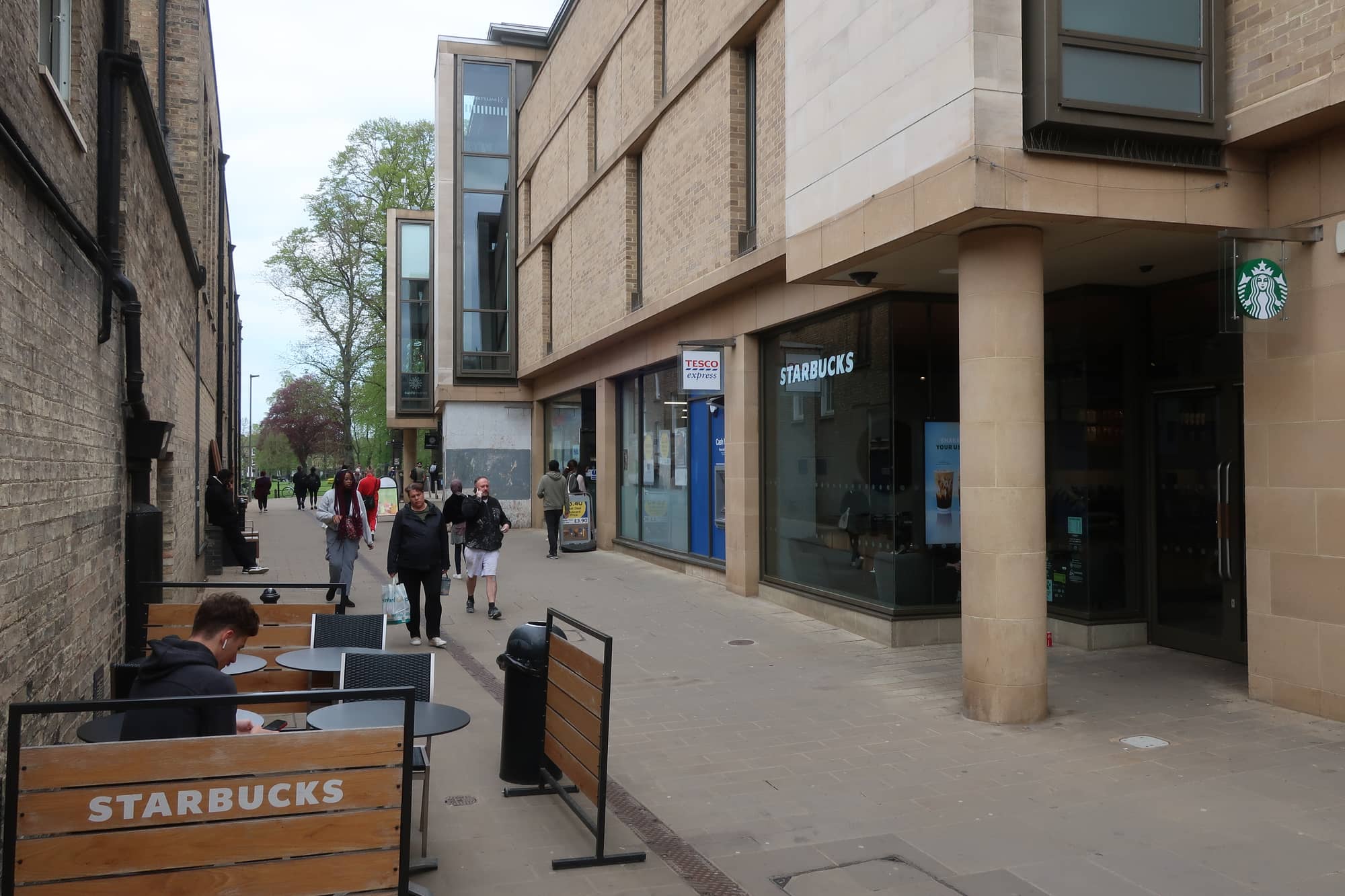Christ's Lane in Cambridge in May 2023, a pedestrian street of shops including a Tesco Express and a Starbucks under a modern stone-clad building, with people walking towards the trees at the far end