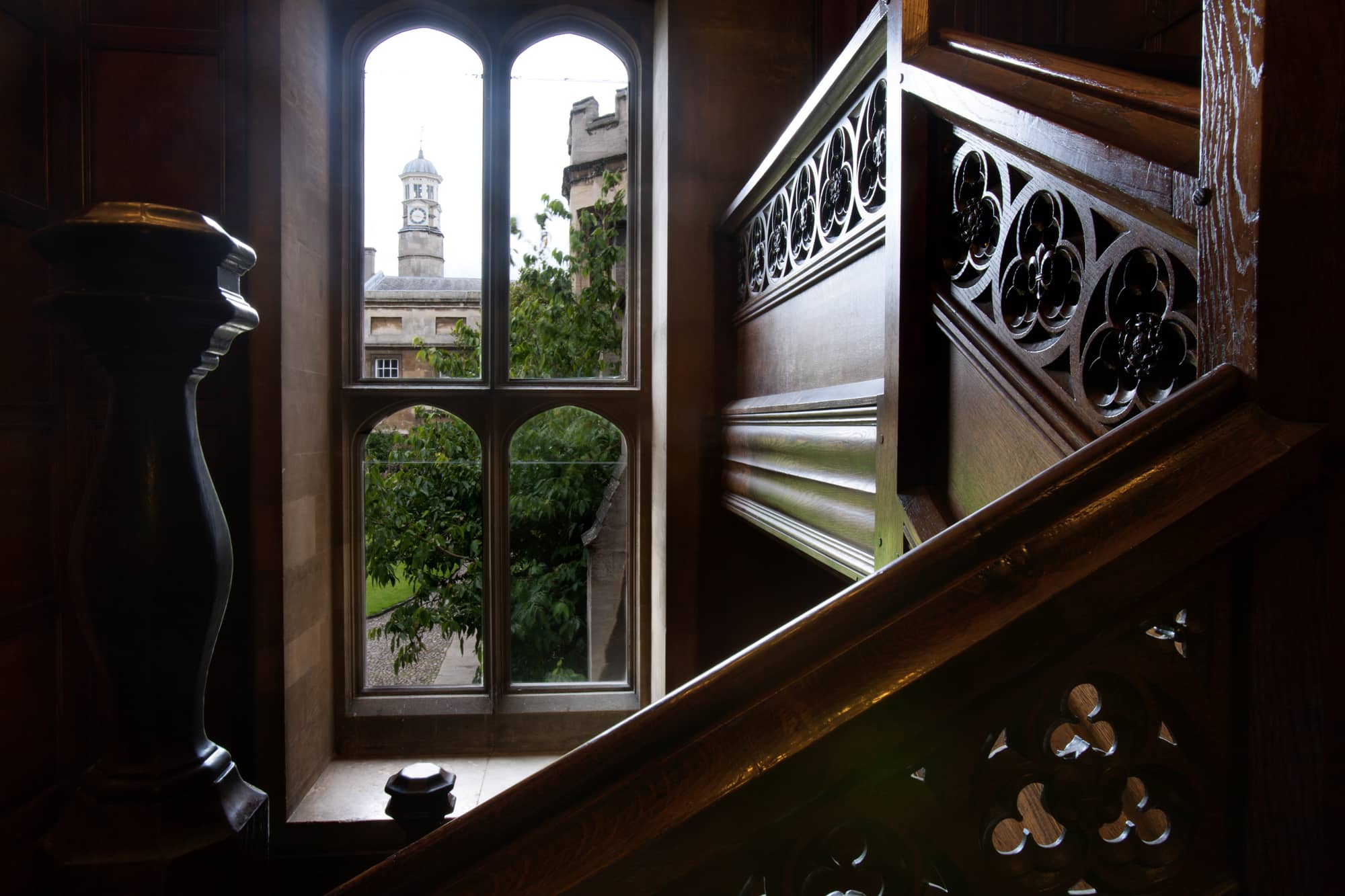 A carved oak staircase with pierced quatrefoil panels inside Christ's College, Cambridge, beside a stone mullioned window looking out to a court and a clock turret