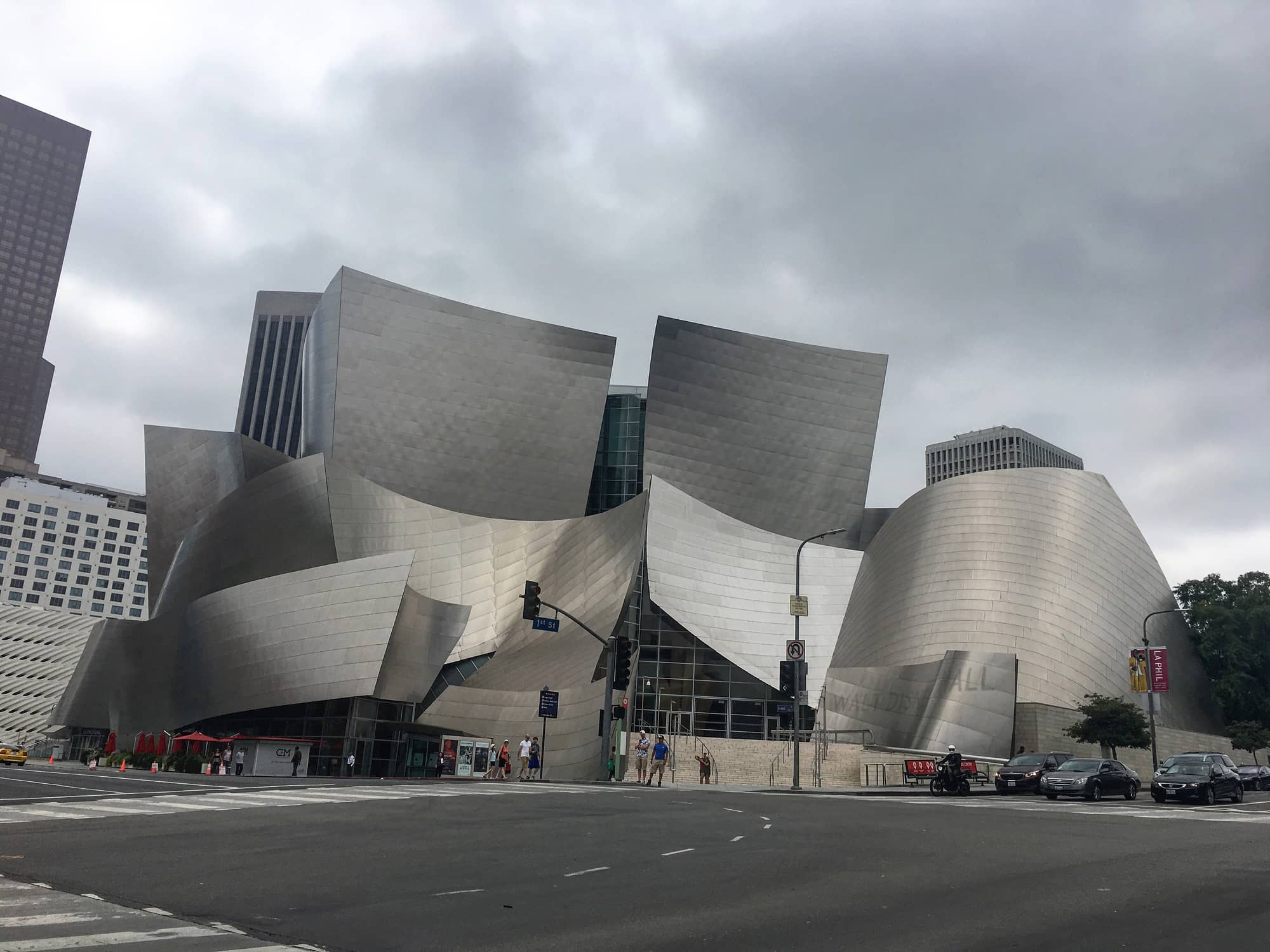The curved stainless-steel sails of Frank Gehry's Walt Disney Concert Hall on Grand Avenue under a cloudy sky, with downtown towers behind