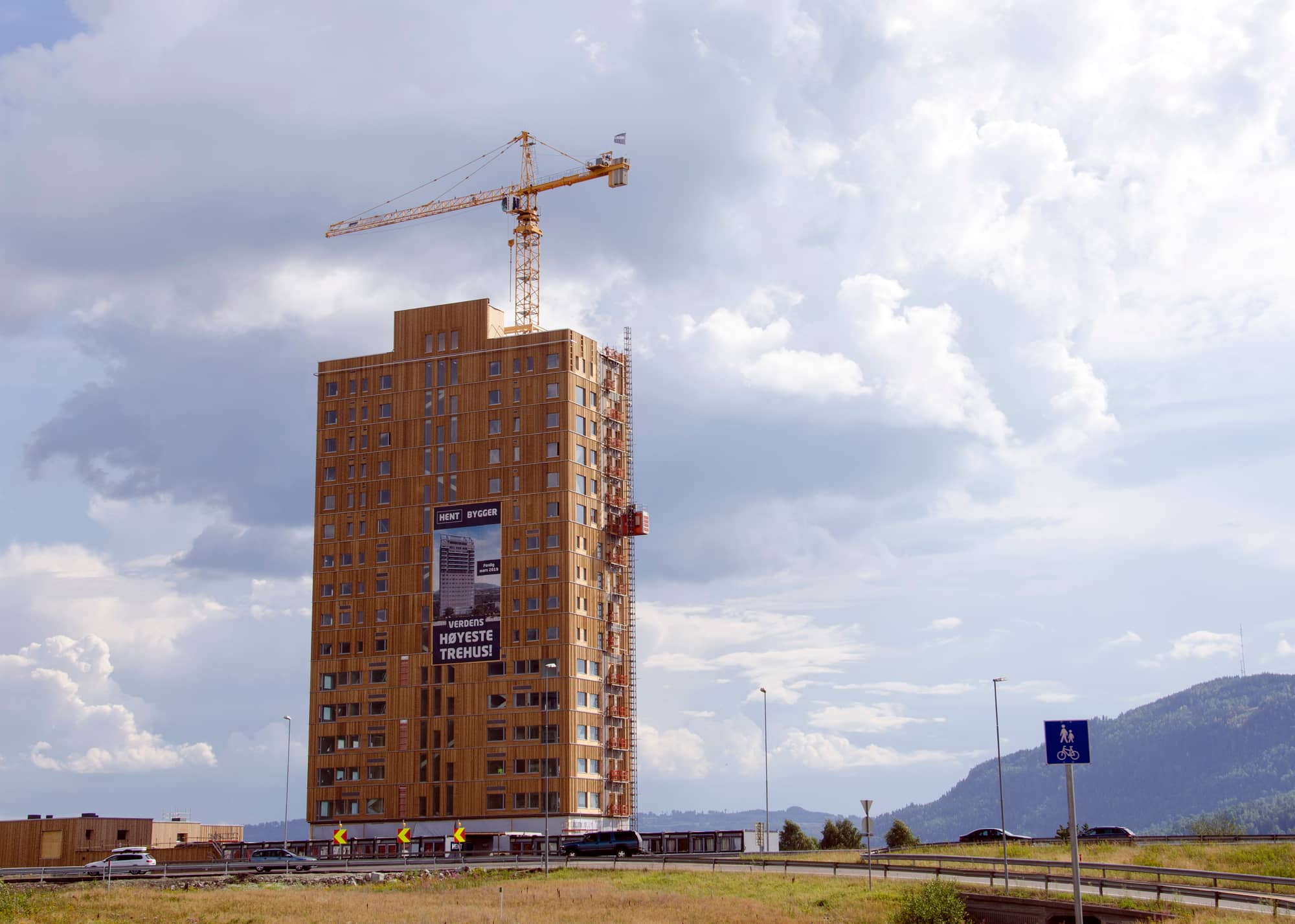 Mjøstårnet, a timber-clad tower in Brumunddal, Norway, under construction with a crane on top and a banner on its facade