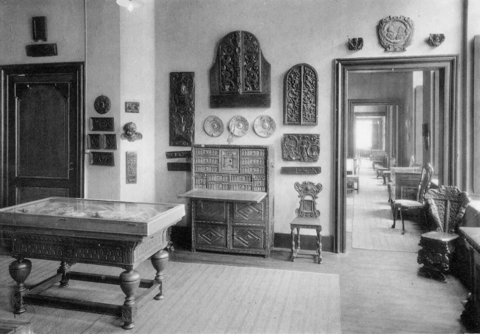 A black-and-white photograph of an early room of Design Museum Gent, with carved furniture, wall plaques and doorways leading through a row of rooms