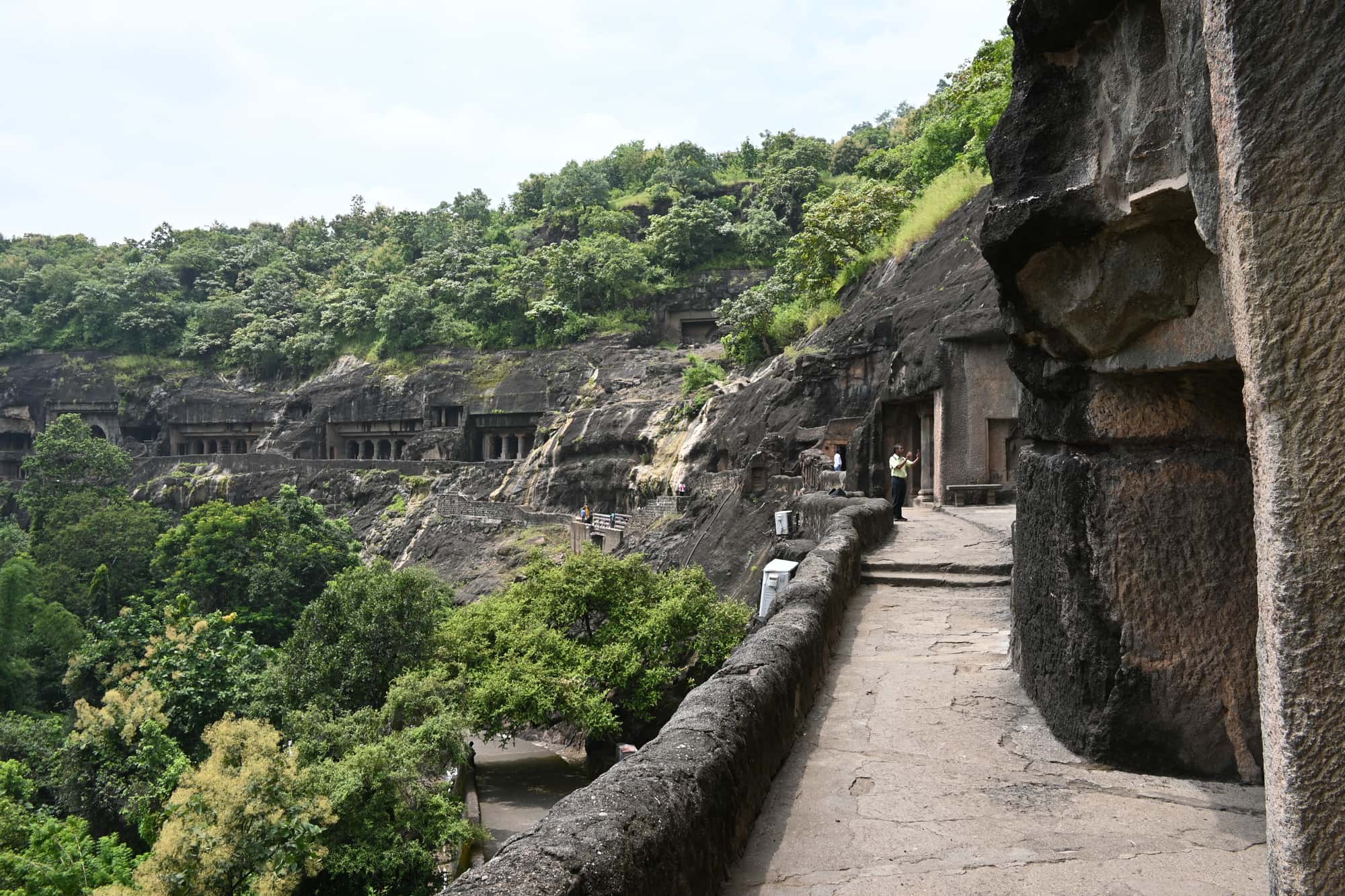 A stone walkway along a cliff of dark basalt at the Ajanta caves in Maharashtra, India, with carved cave entrances and pillared porches visible across a wooded gorge