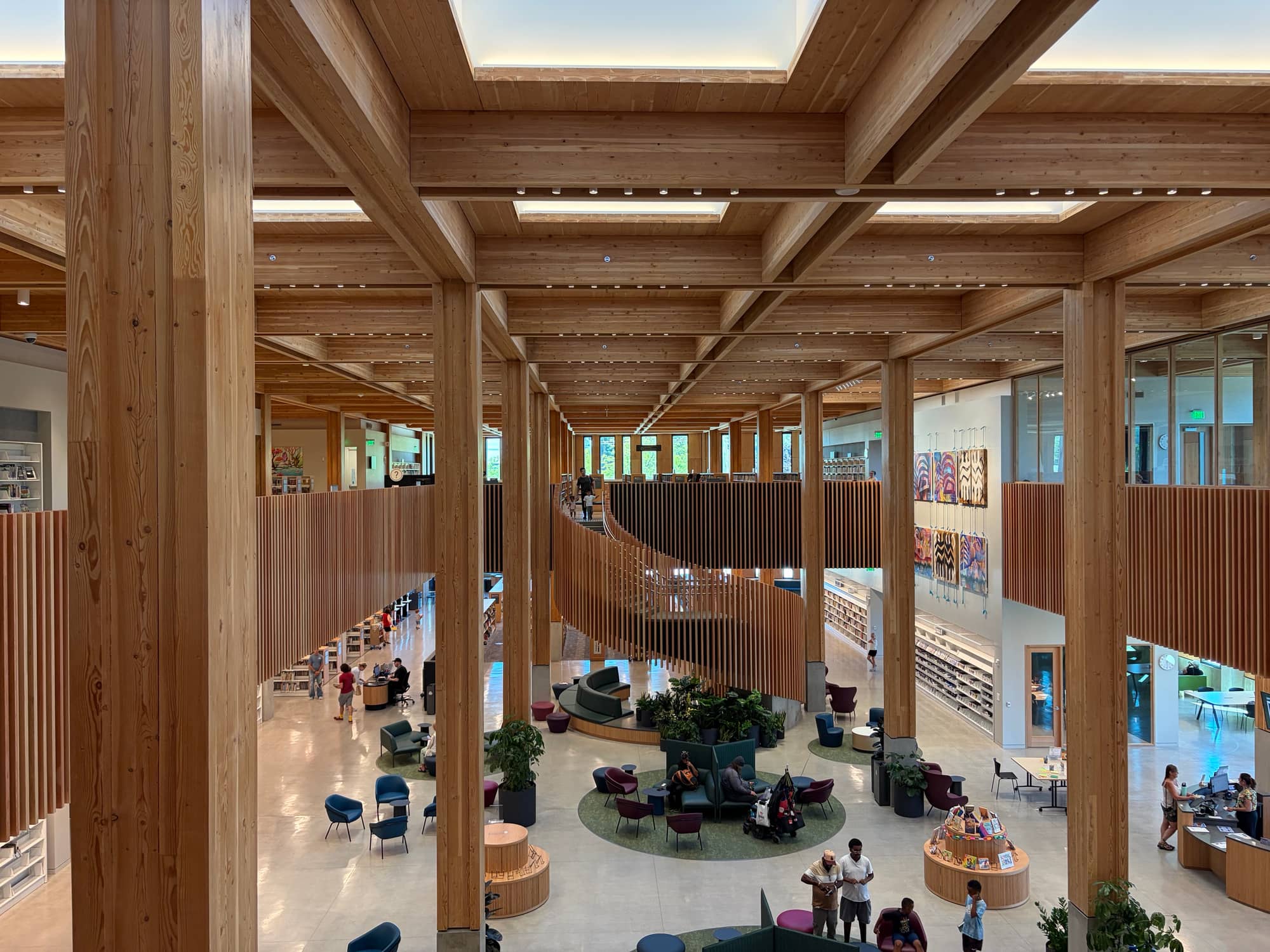 Inside the East County Library: a double-height atrium framed by a grid of exposed glulam columns and beams under skylights, with a spiral staircase wrapped in timber slats and reading chairs on a pale concrete floor