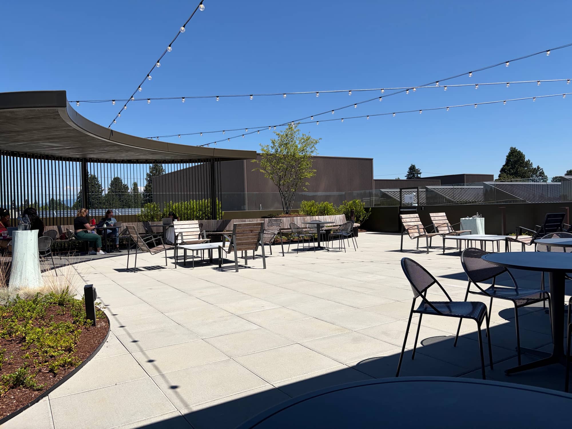 The rooftop terrace of the East County Library in Gresham: paving, tables and wooden lounge chairs under strings of lights, beside a curved canopy on slender posts, with solar panels and trees beyond