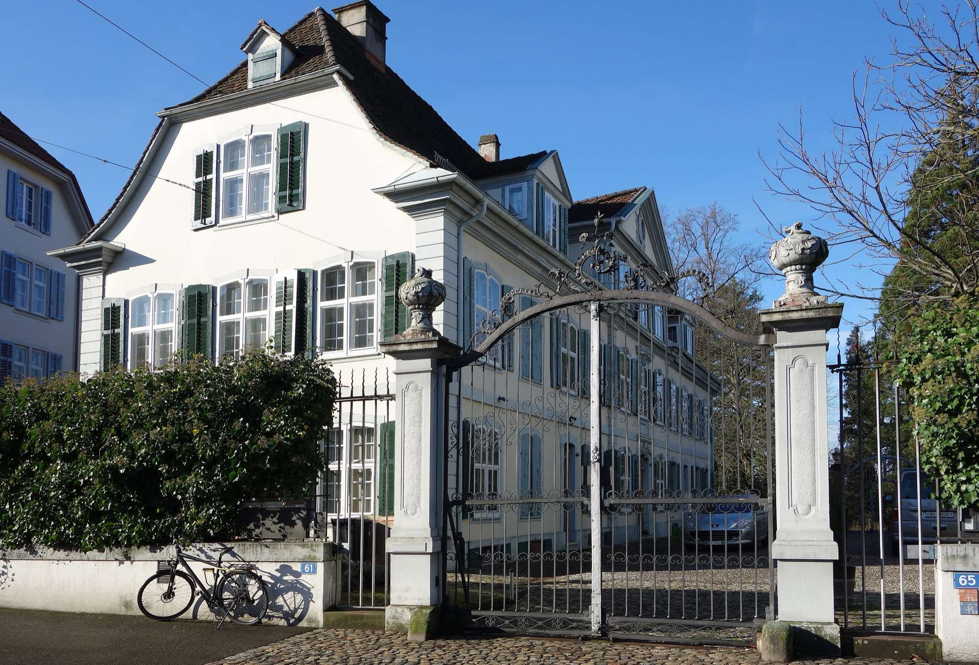 A white three-storey villa with green shutters behind an ornate iron gate between urn-topped stone pillars on Baselstrasse in Riehen