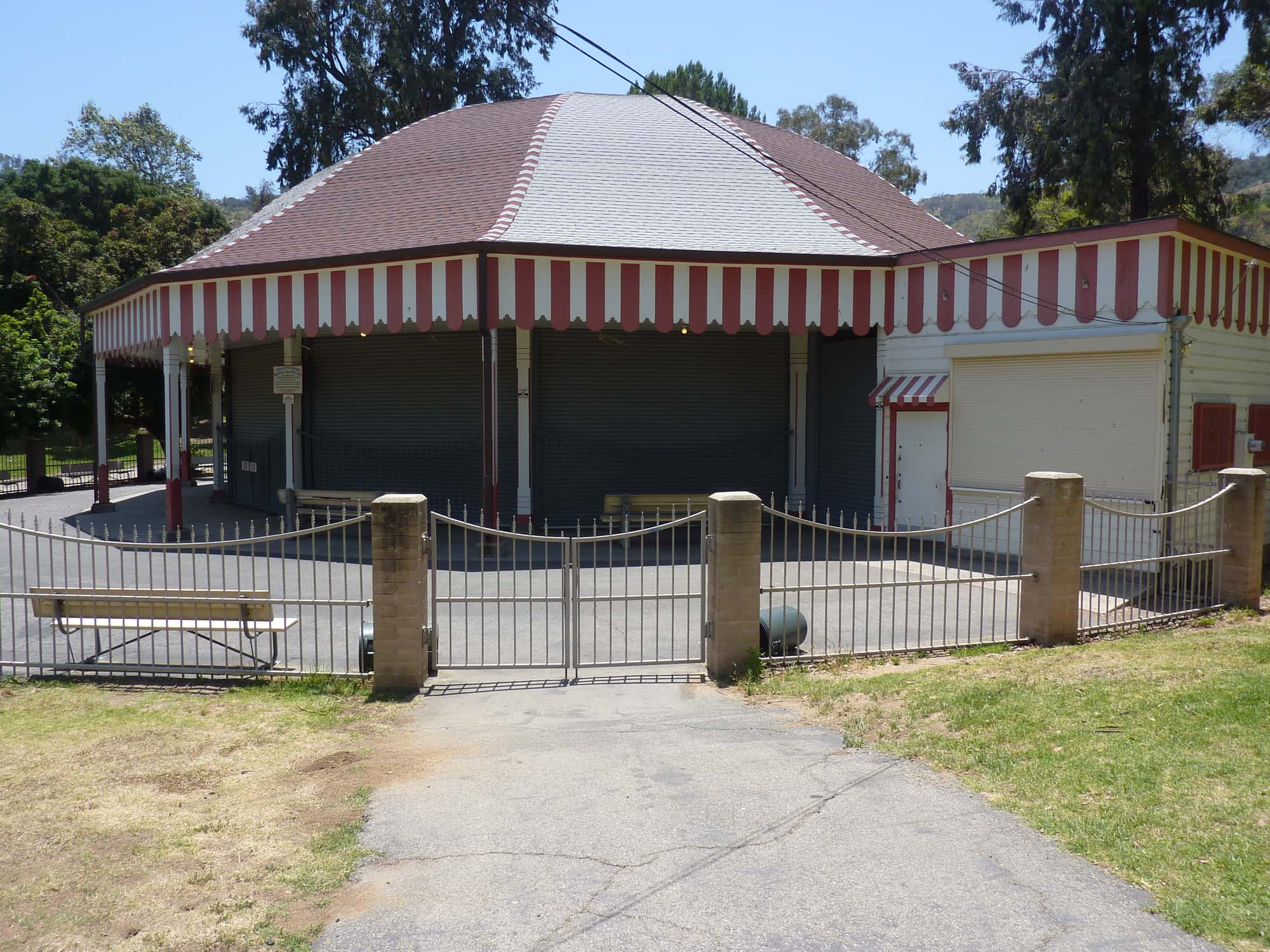 The Griffith Park merry-go-round pavilion seen from the north in 2010, its striped roof and red-and-white valance over closed shutters, with a gated path and a bench in front