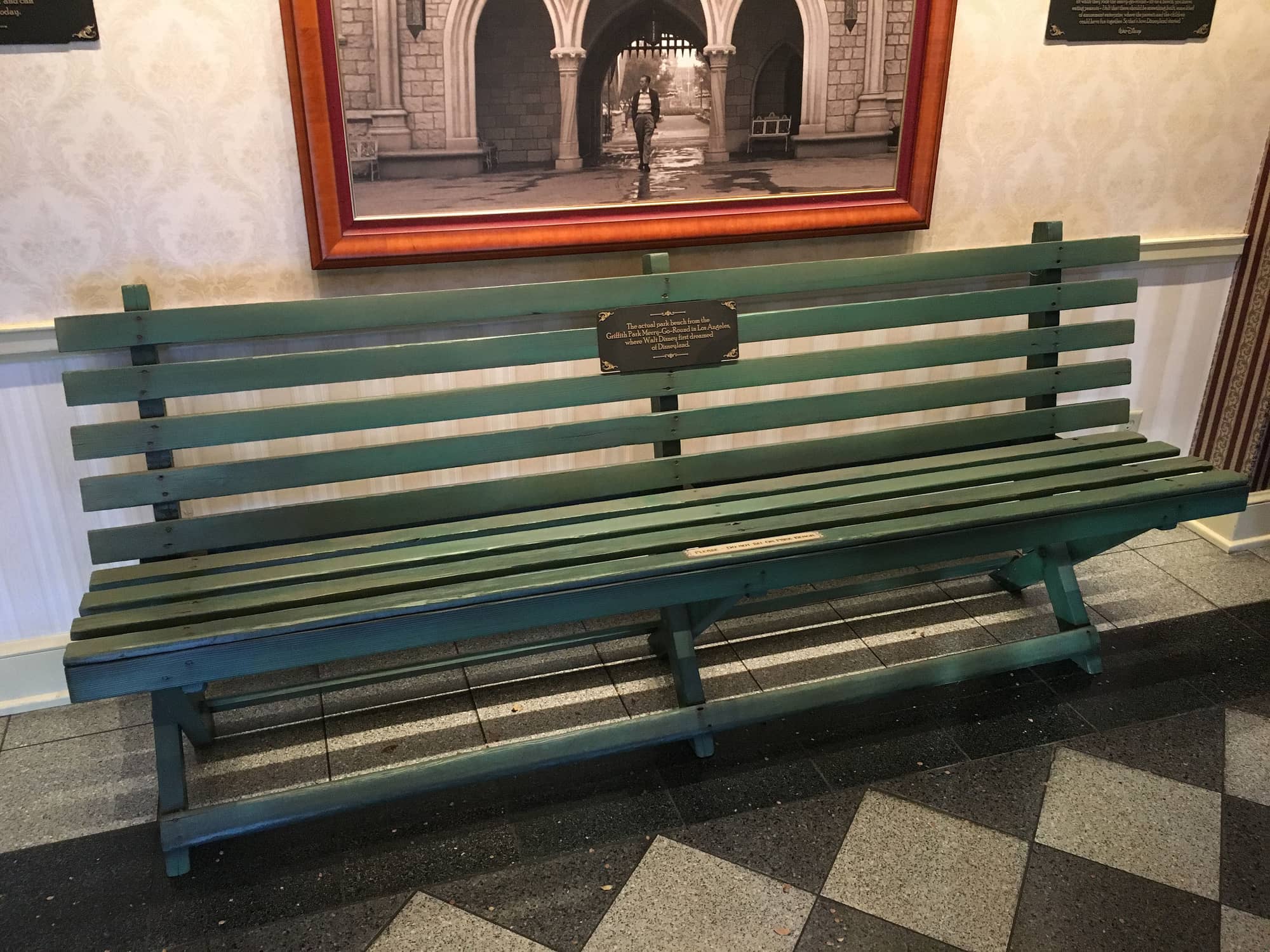 A green-painted wooden park bench under a framed photograph of Walt Disney, with a plaque saying it is the actual park bench from the Griffith Park Merry-Go-Round where Walt Disney first dreamed of Disneyland