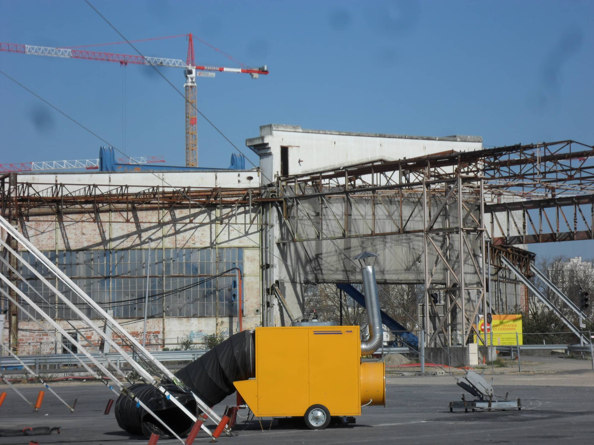 The steel frame of the old Renault factory access portal on the Île Seguin in March 2014, with a brick and glass hall behind, a yellow site heater in front and a crane overhead