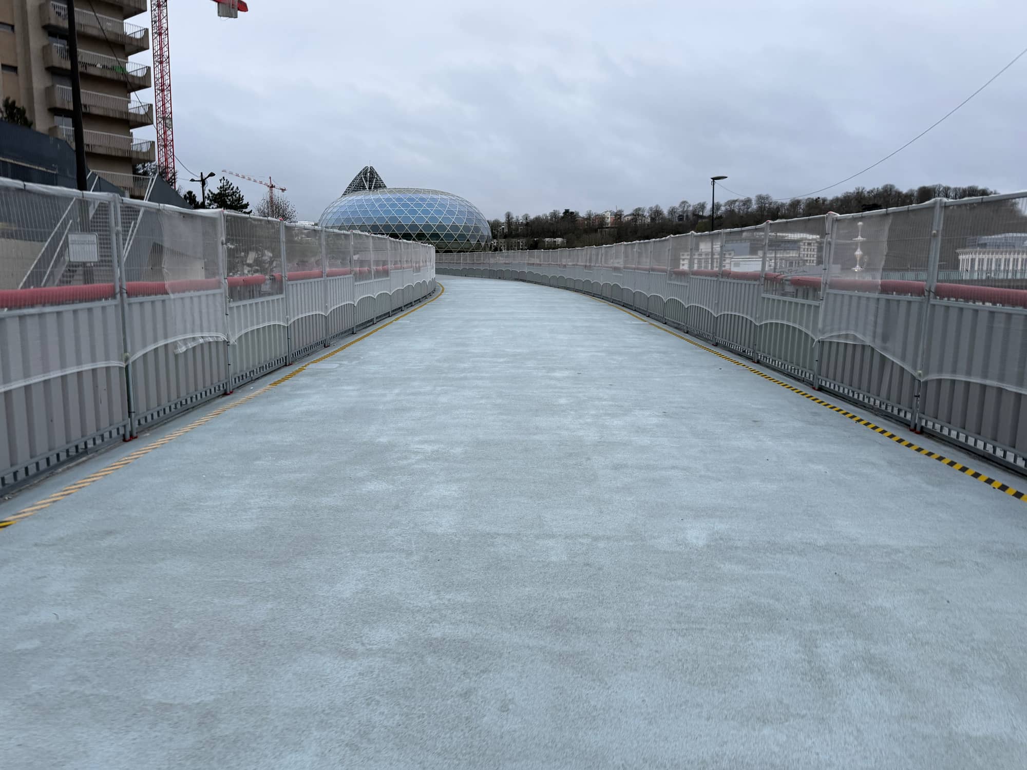 A new footbridge deck with white mesh railings leading onto the Île Seguin in February 2026, with the glass sphere of La Seine Musicale ahead and a crane to the left