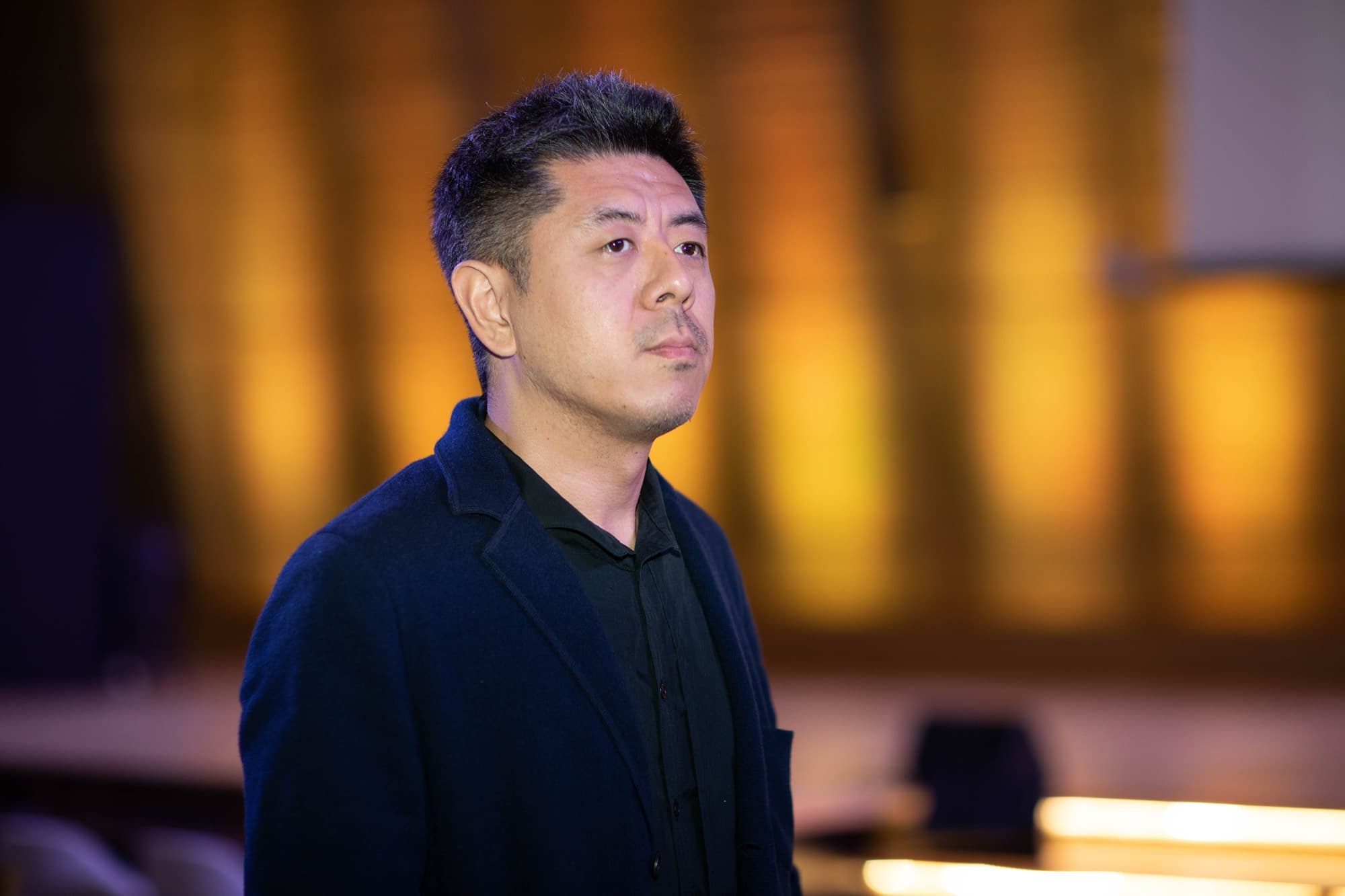 Waist-up portrait of Ma Yansong in a dark navy jacket against a blurred amber background