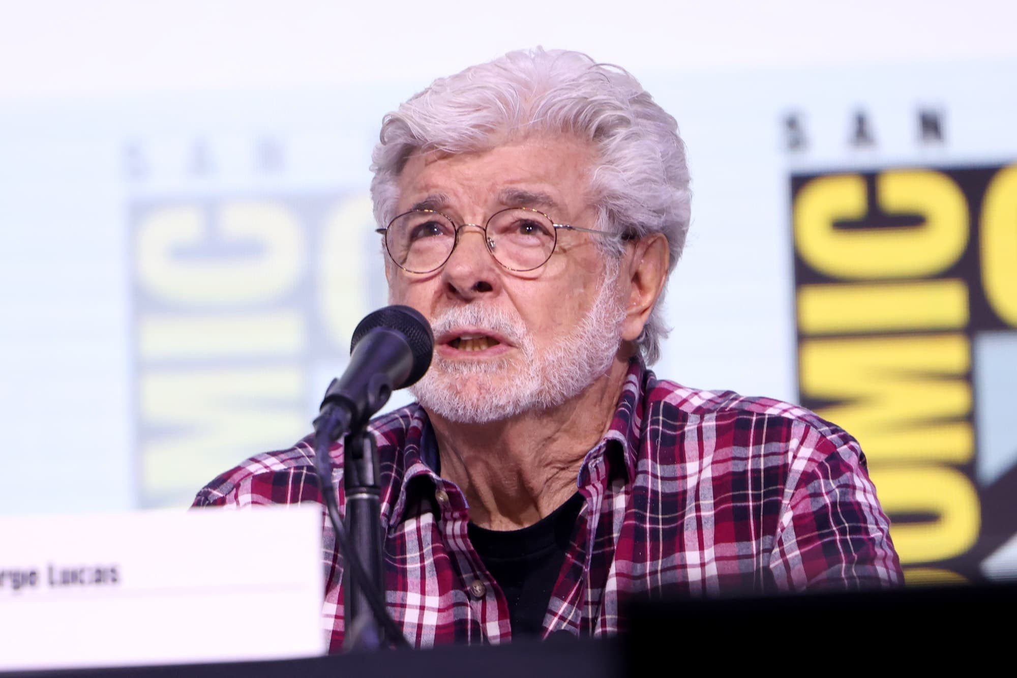 George Lucas, white-haired and bearded, in a red plaid shirt, speaking into a microphone at San Diego Comic-Con