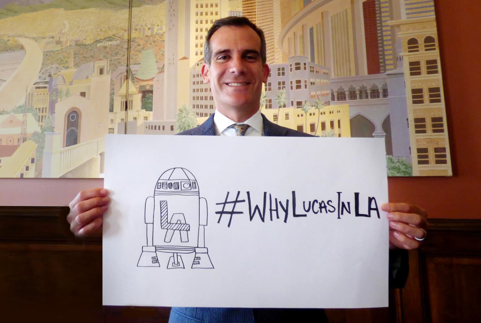 Eric Garcetti smiling and holding a hand-drawn sign of an R2-D2-like robot marked LA and the hashtag WhyLucasInLA in front of a painted mural