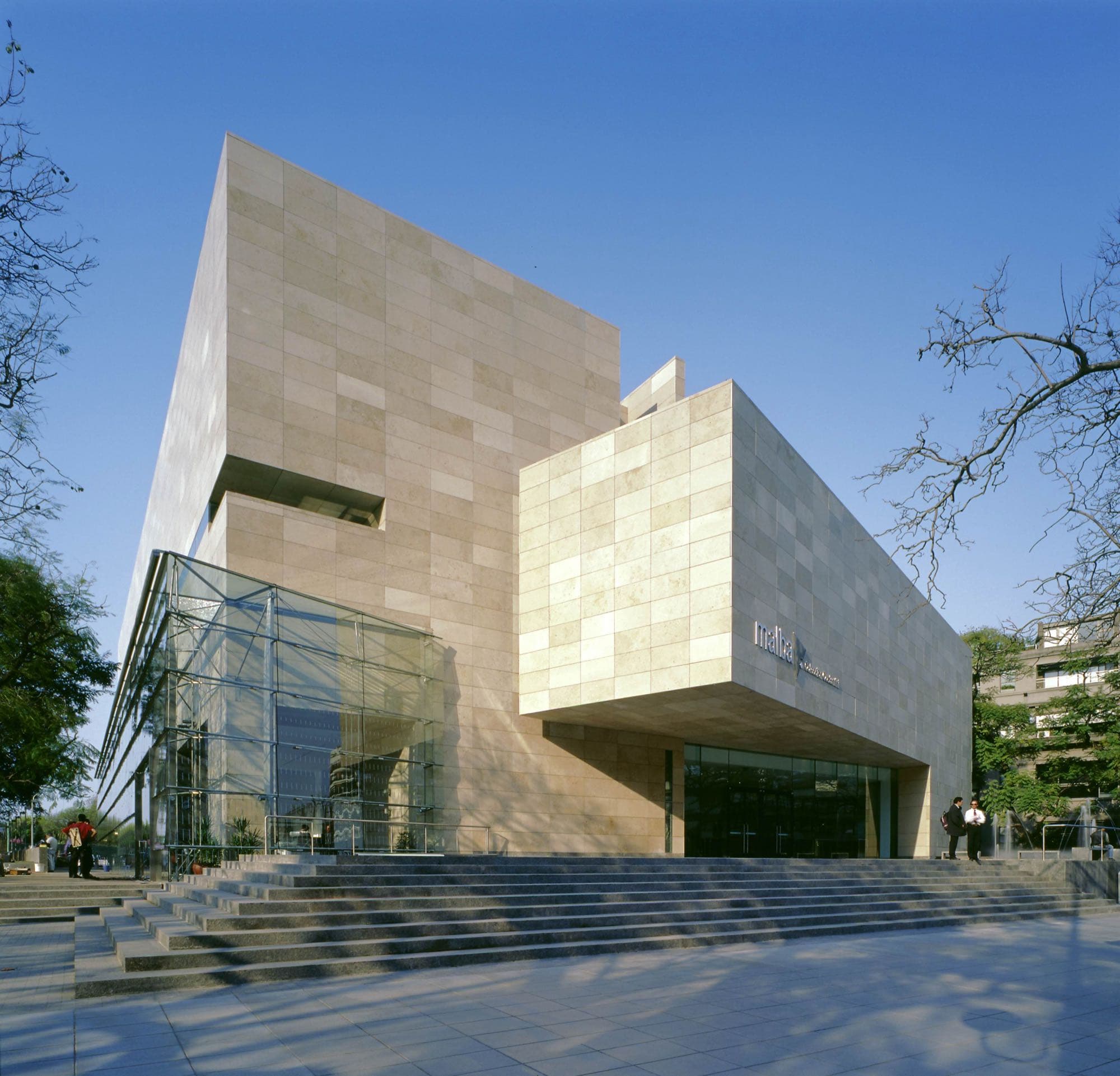 Corner view of Malba's stacked limestone prisms, a tall glass box at left and a cantilevered block over a recessed glazed entrance above wide steps