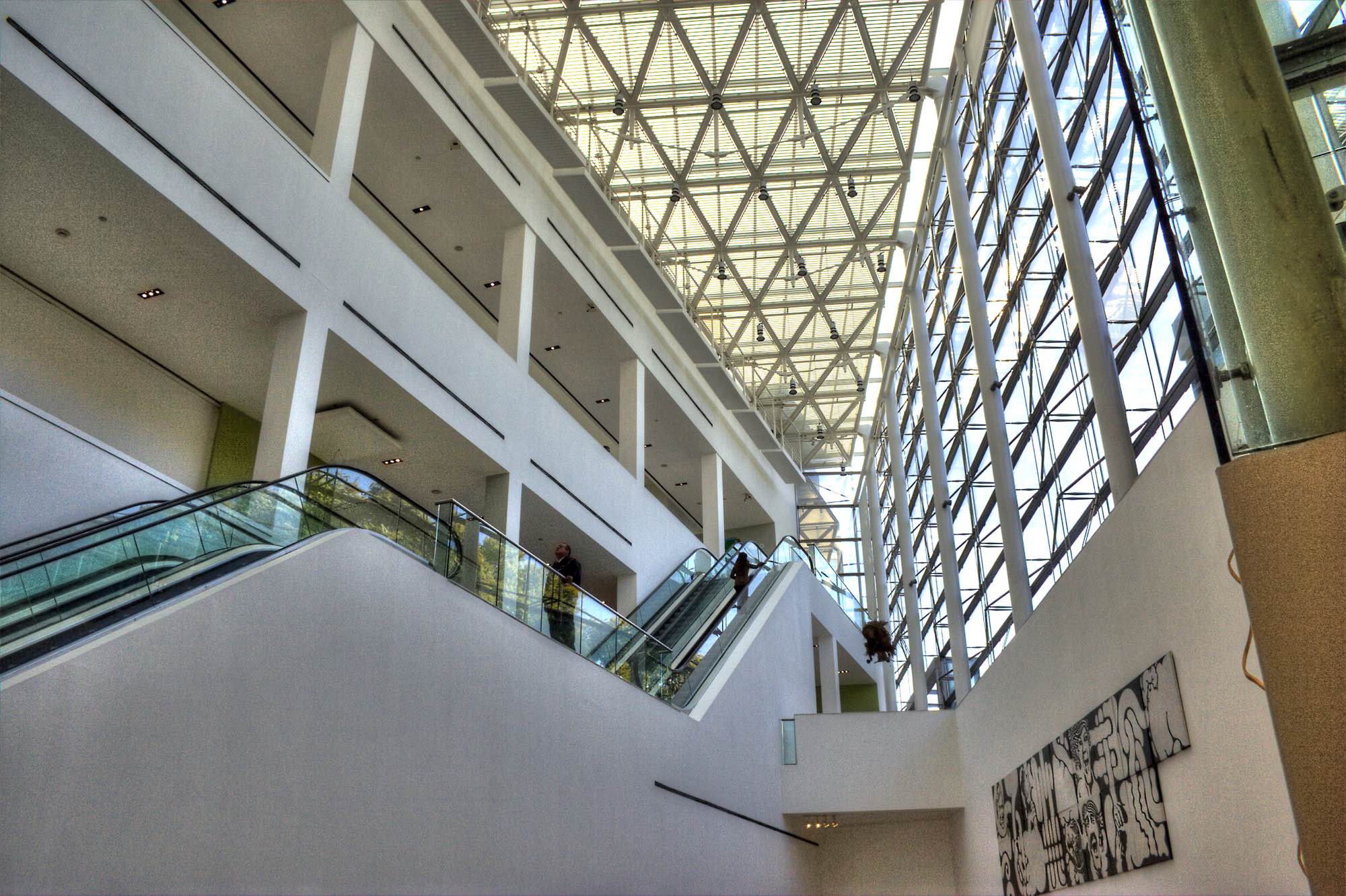 Malba's full-height atrium: escalators with glass balustrades rise between white gallery floors under a triangulated skylight