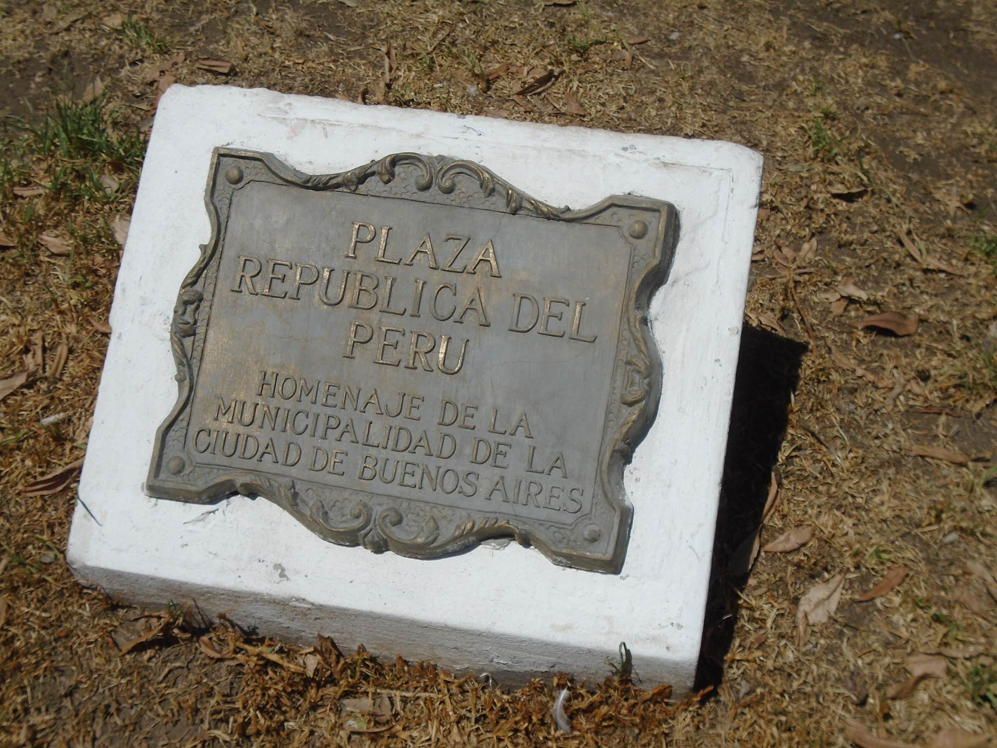 A bronze plaque reading Plaza República del Perú on a white plinth in dry grass