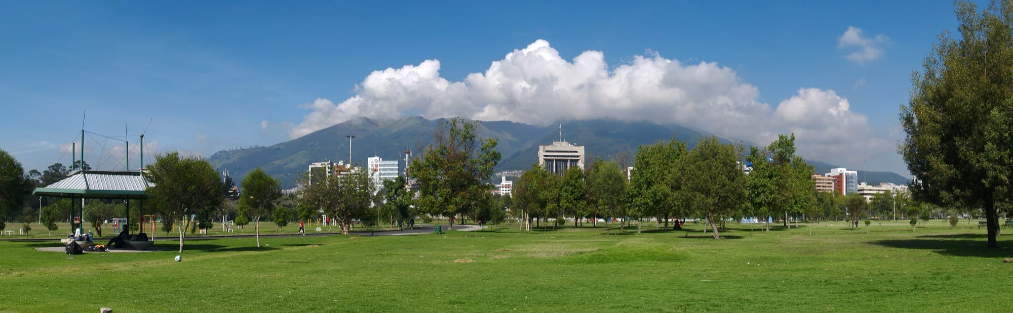Wide panorama of the lawns and trees of Parque La Carolina in Quito, with a pavilion at left and office towers along the park edge below a cloud-capped mountain range