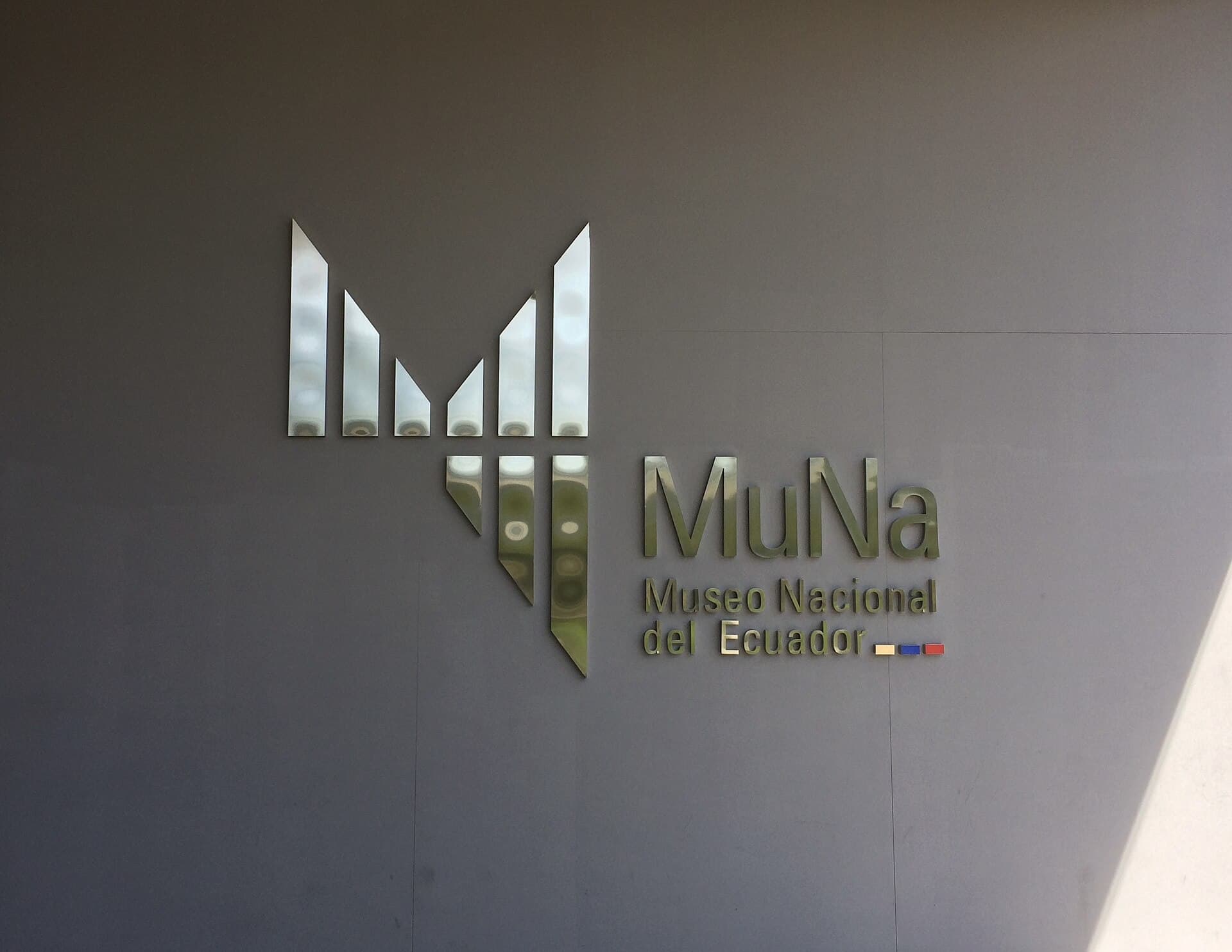 Polished-metal MuNa logo and the words Museo Nacional del Ecuador on a grey wall at the museum's current entrance