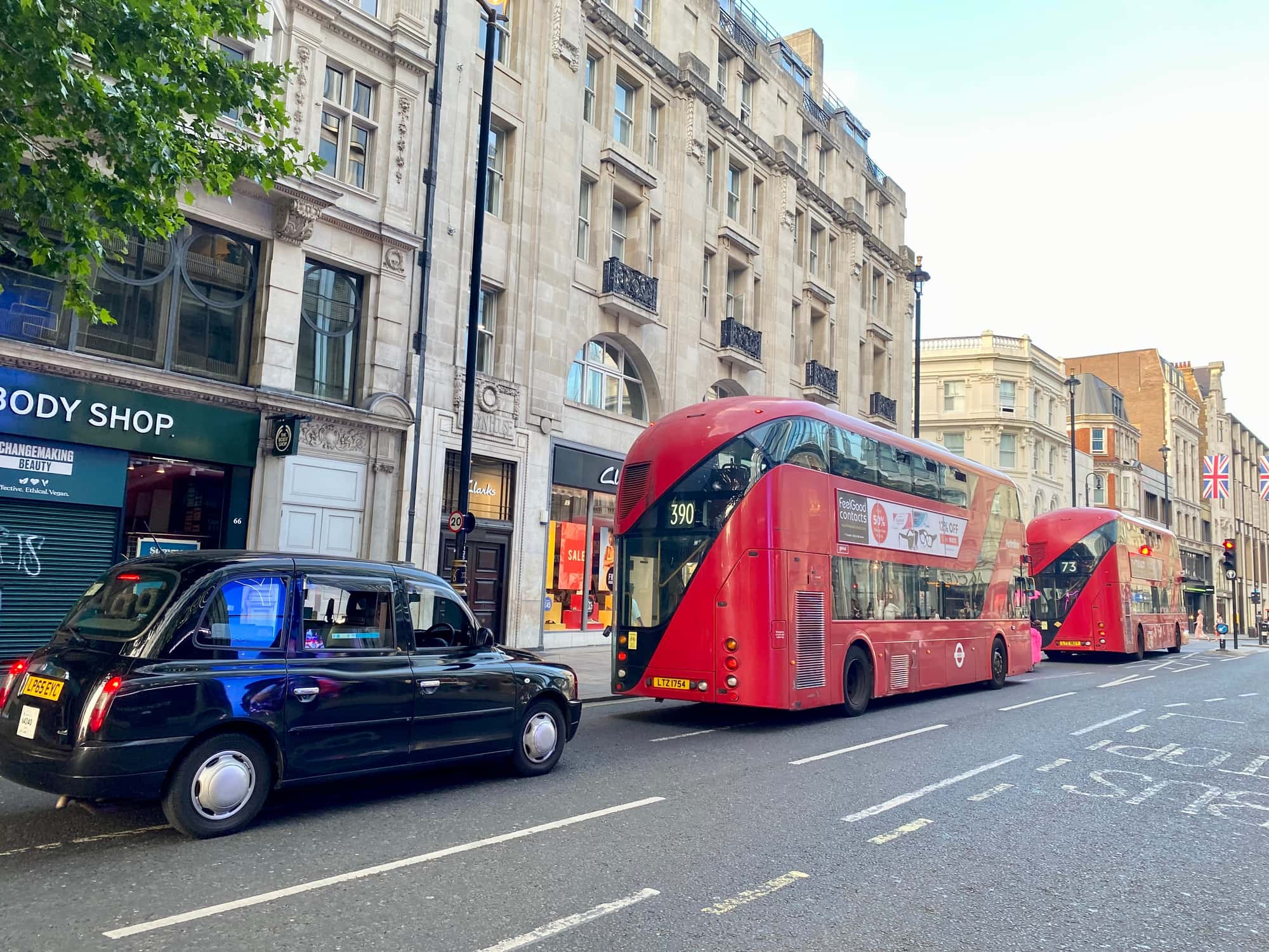 A black taxi and two red double-decker buses on routes 390 and 73 driving along Oxford Street past stone-fronted shops in July 2025
