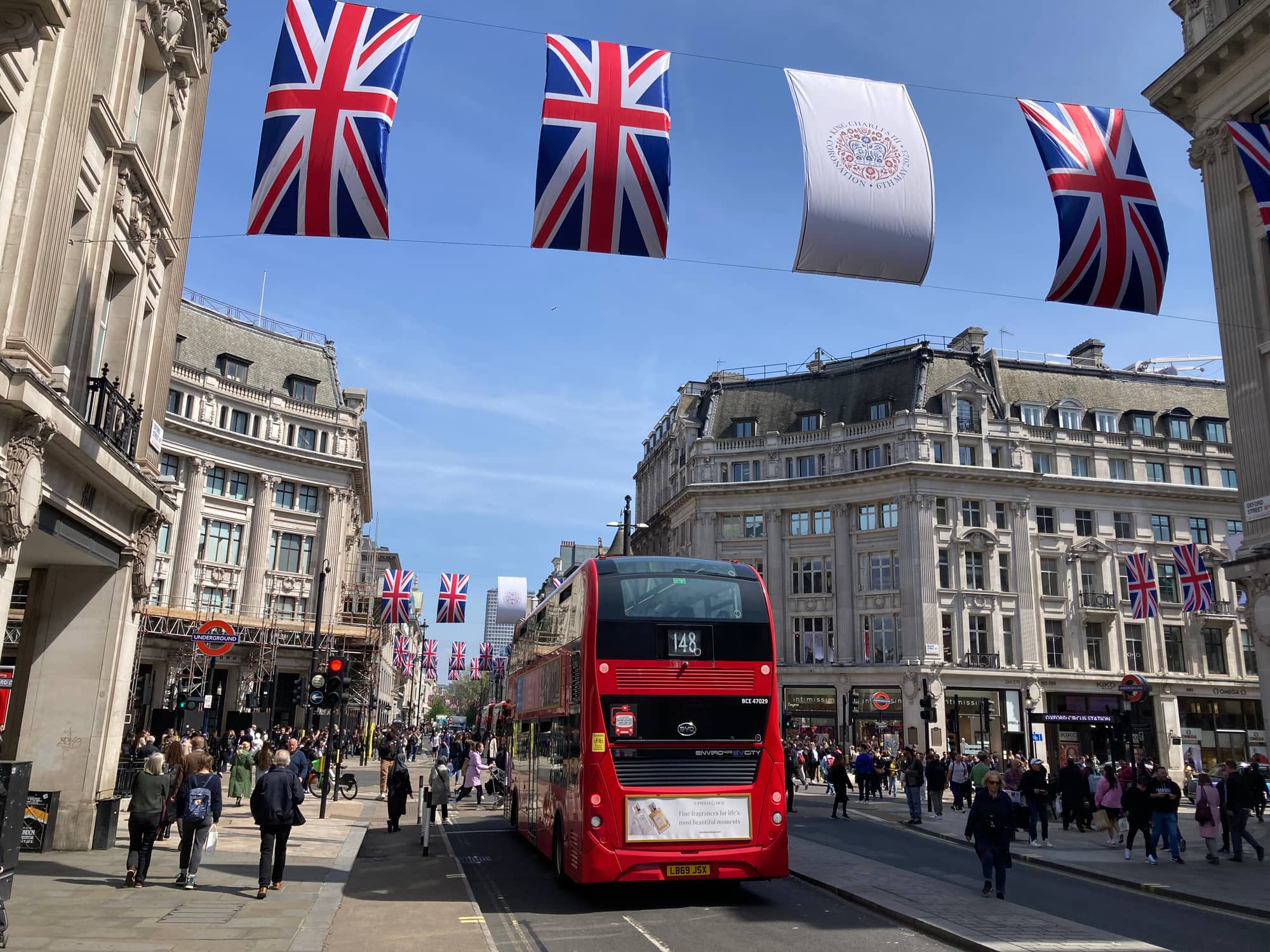 Oxford Circus in May 2023, with a red bus, crowded pavements, an Underground entrance and Union flags strung across the junction between curved stone buildings