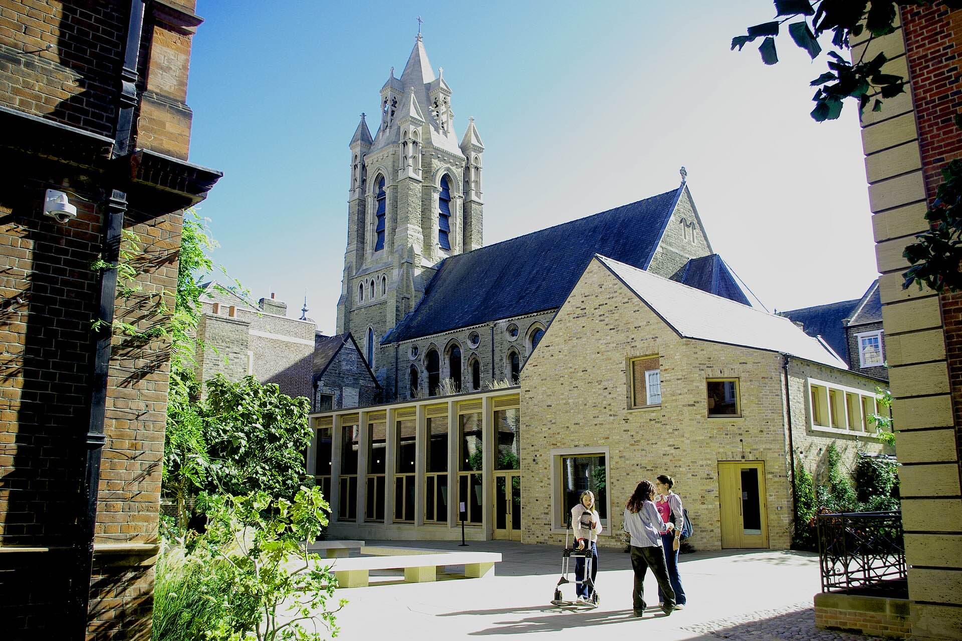 A sunlit paved court at Pembroke Mill Lane with a Gothic Revival stone church tower fronted by a single-storey timber-framed glazed foyer