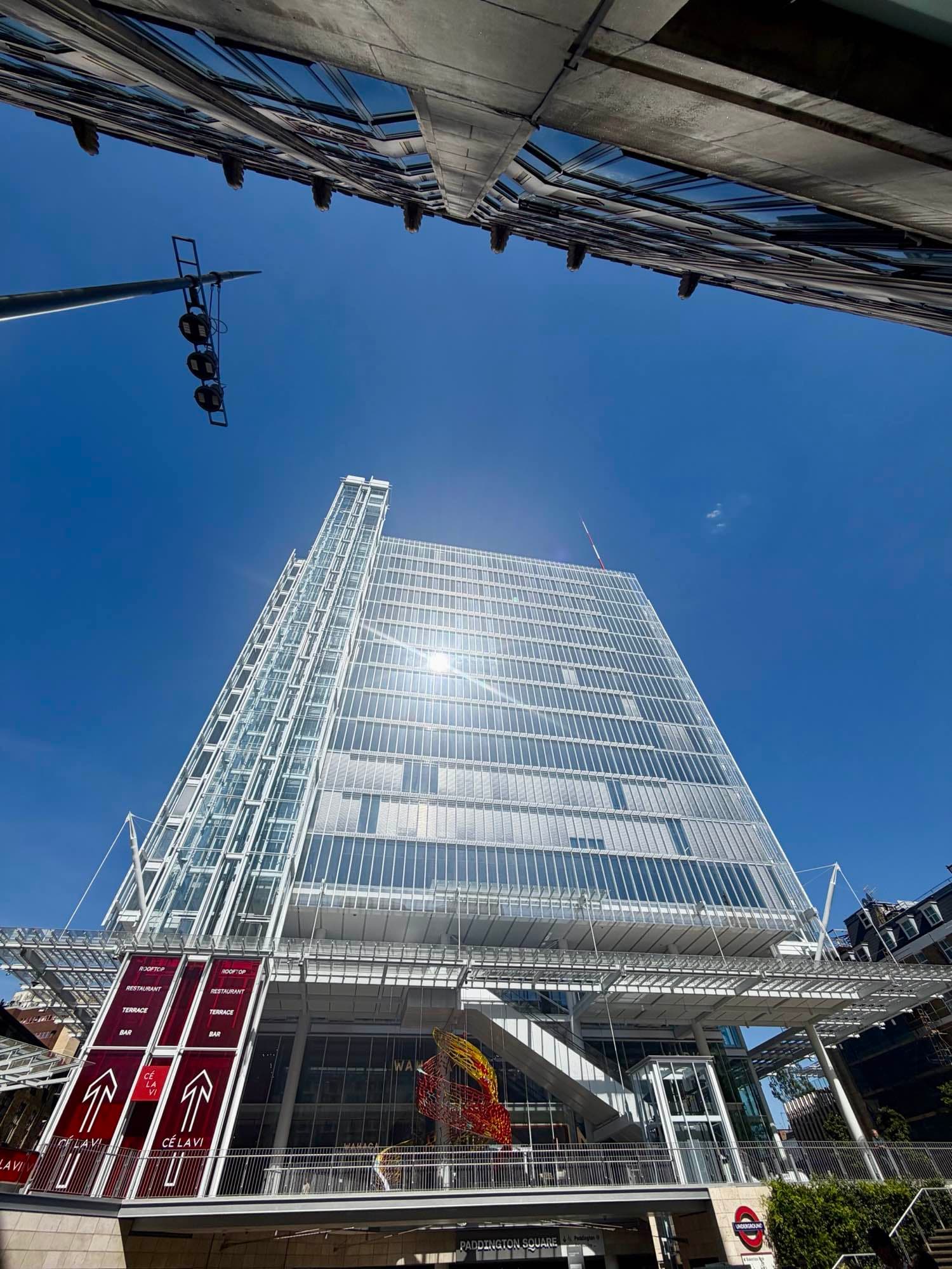 Looking up at Paddington Square's glass-clad block above a white steel canopy, with a glazed lift tower and an Underground roundel at the base