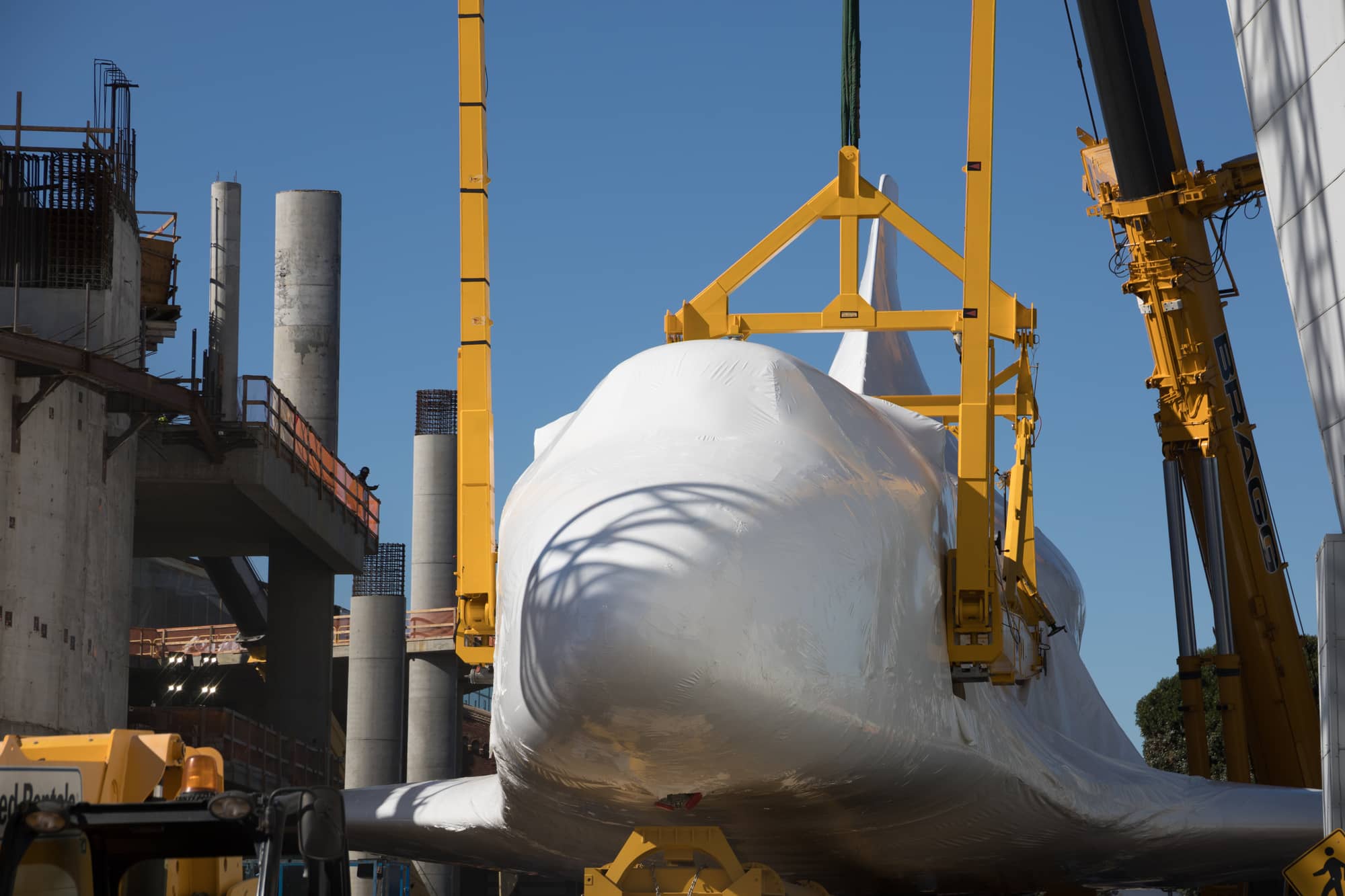 Space Shuttle Endeavour wrapped in white protective film, held in a yellow lifting frame between cranes, beside concrete columns of the Samuel Oschin Air and Space Center in January 2024