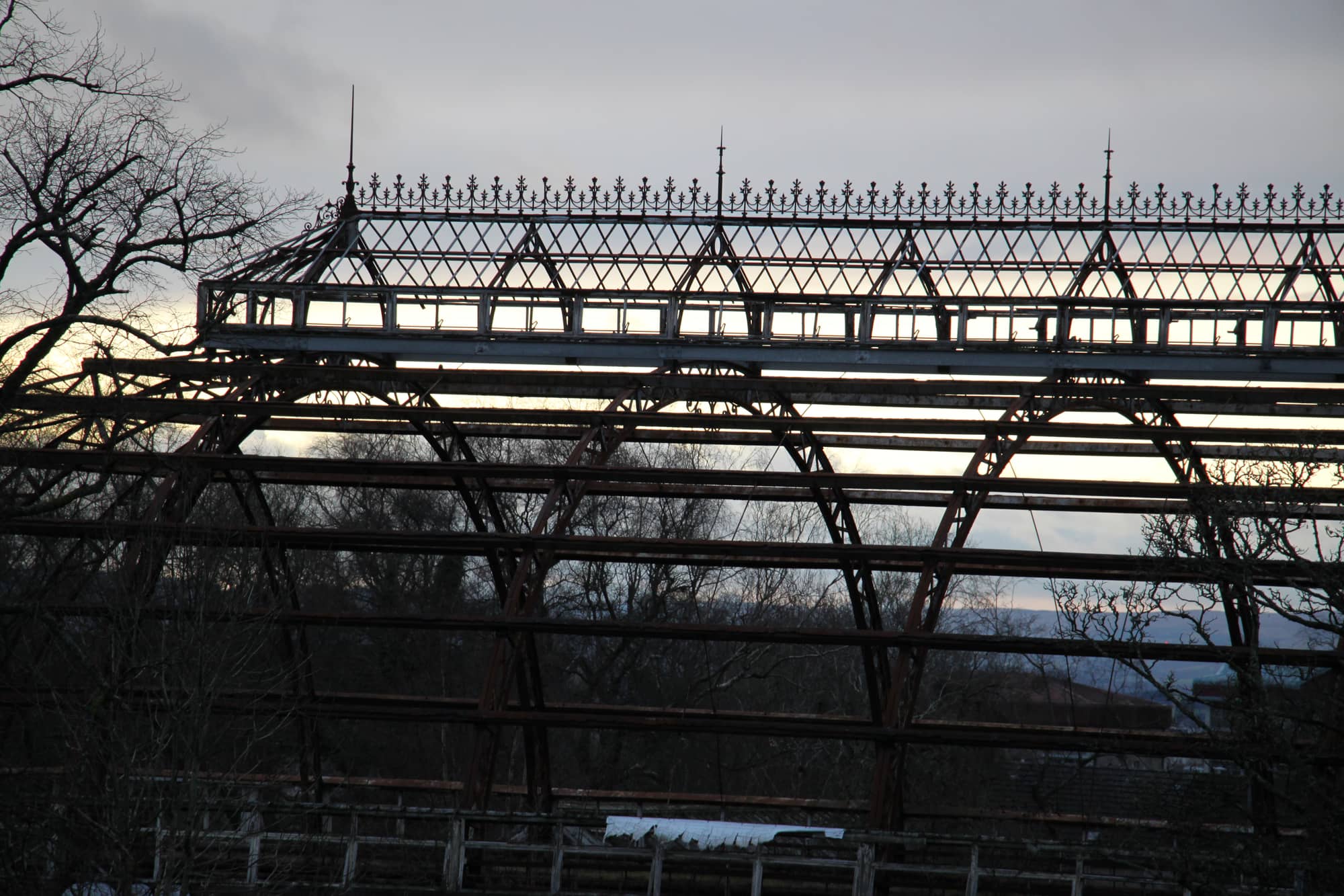 The ridge of the Springburn Winter Gardens at dusk in December 2024: a long lantern of iron glazing bars topped with decorative cresting and finials, above the bare arches of the main hall