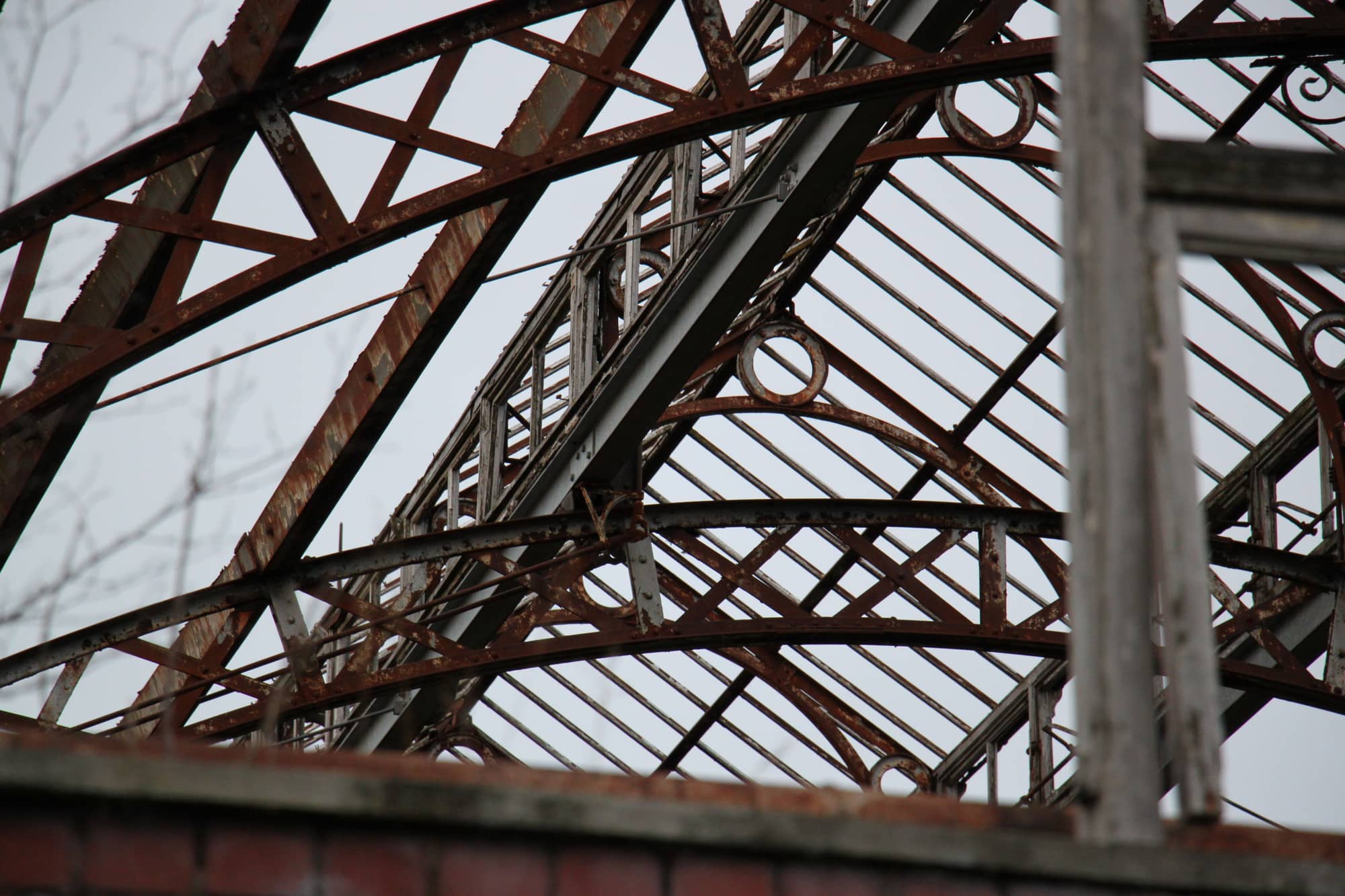 Close-up of the rusted lattice trusses and curved glazing bars of the Springburn Winter Gardens roof against a pale sky