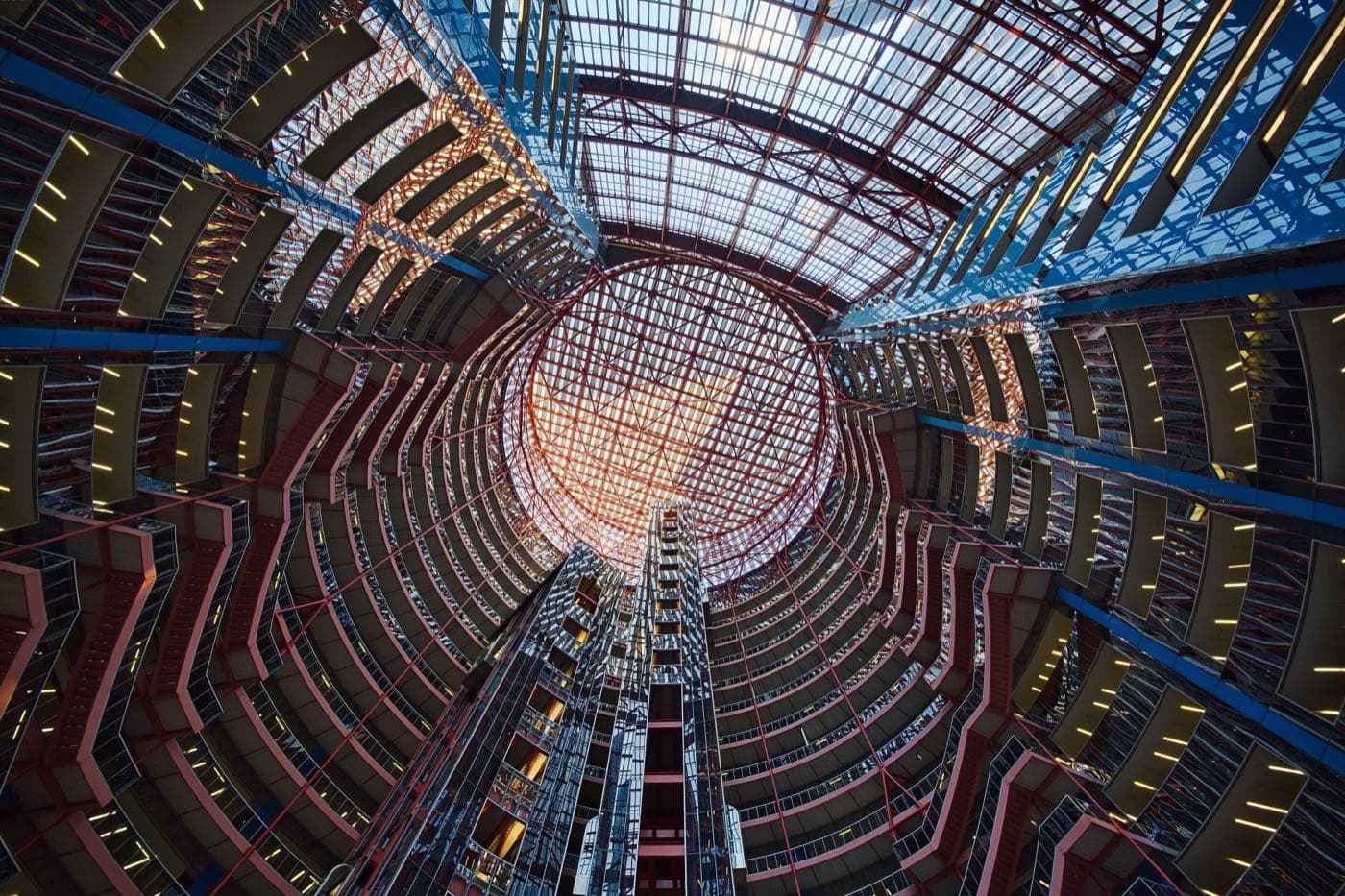 Looking up the Thompson Center atrium: curved balconies trimmed in salmon and blue rise to a round glass skylight, with a glazed elevator tower at the centre