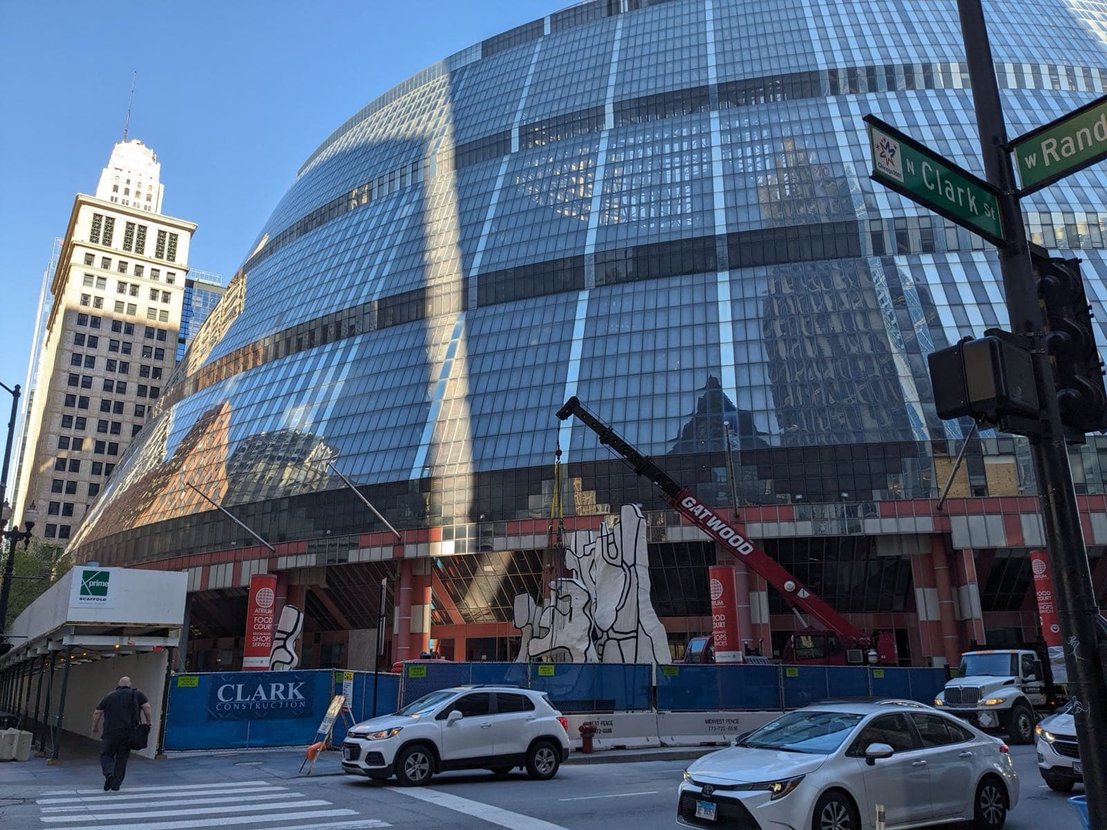 The Thompson Center's reflective glass corner above blue construction hoarding, with a red crane beside the Dubuffet sculpture at Clark and Randolph