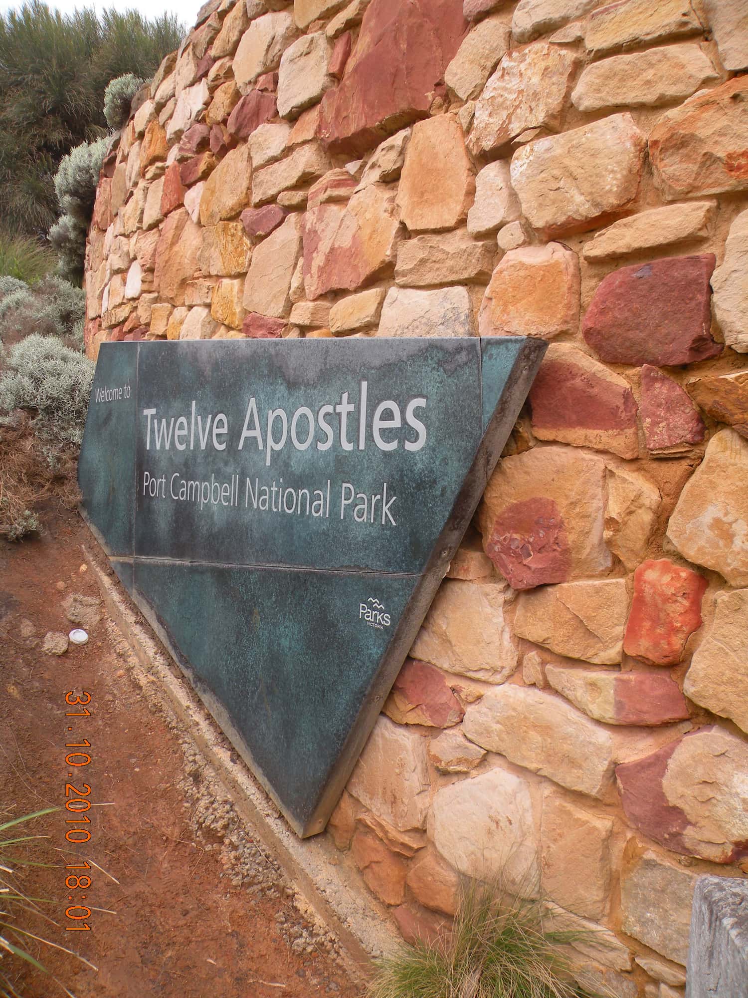 A curved dry-stone wall of red and ochre sandstone with a patinated bronze sign reading Welcome to Twelve Apostles, Port Campbell National Park, photographed in October 2010