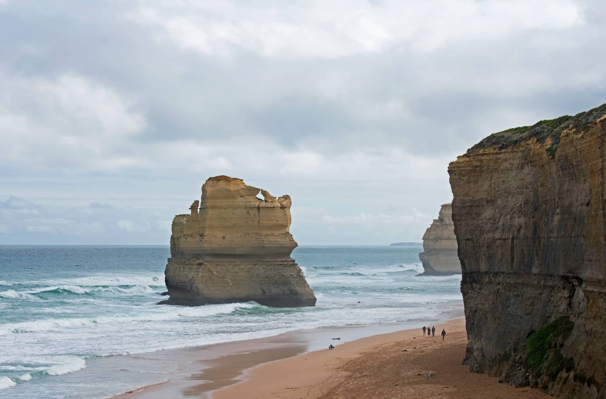 The Twelve Apostles limestone stacks seen from the Gibson Steps at beach level, with waves breaking around them under a pale sky