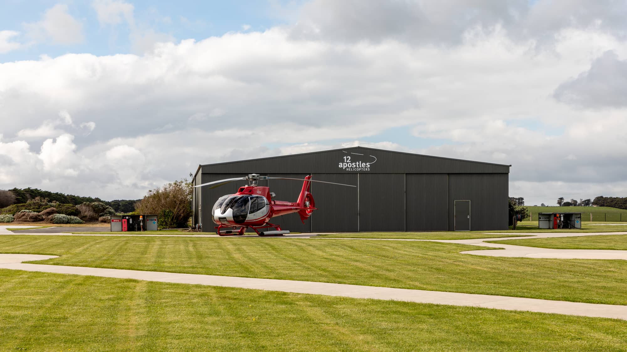 A red and silver sightseeing helicopter parked on a pad in front of a dark corrugated hangar marked 12 Apostles Helicopters, near Port Campbell National Park