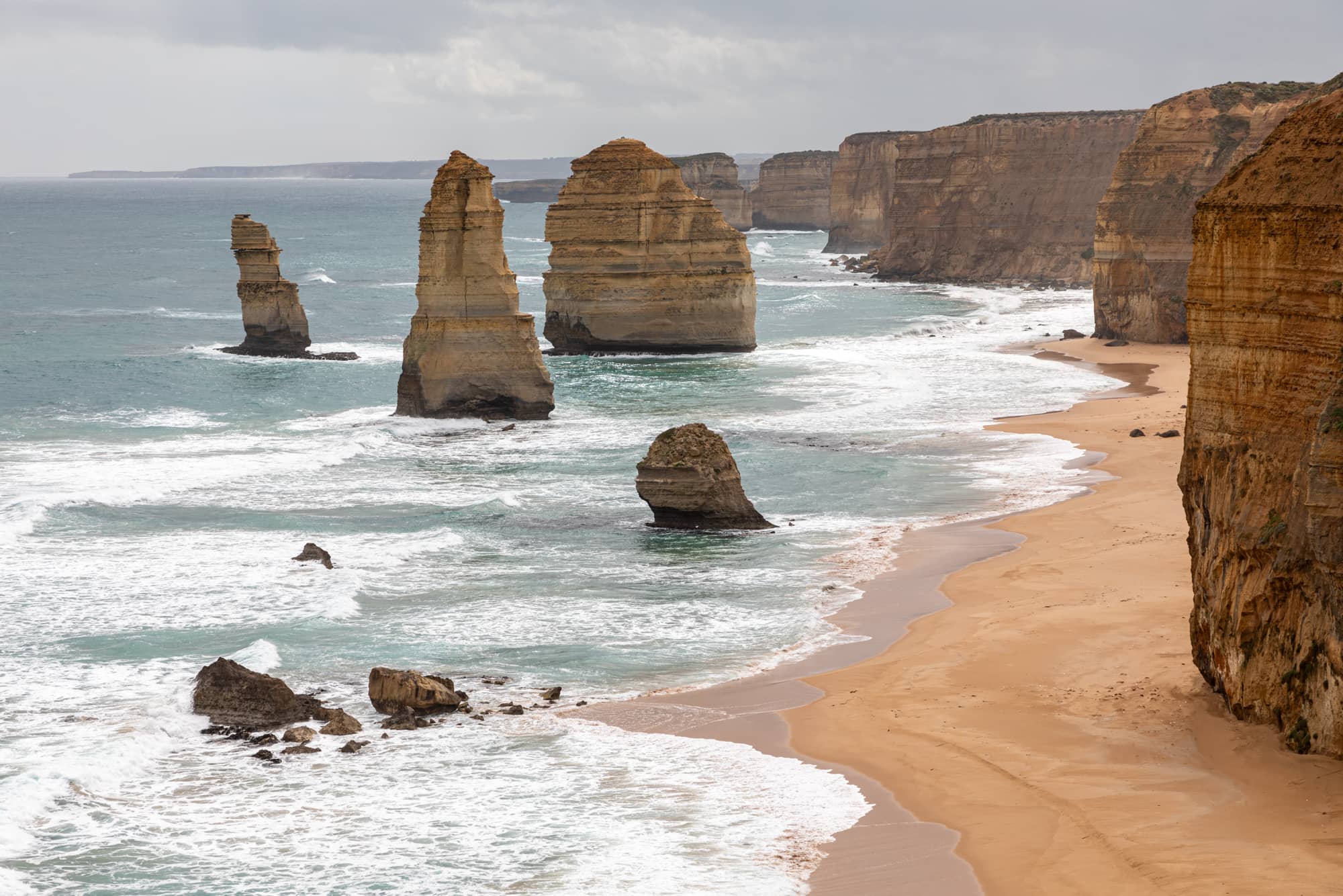 Limestone stacks of the Twelve Apostles standing in surf below sandstone-coloured cliffs and a beach in Port Campbell National Park, the coast the new Twelve Apostles visitor centre serves