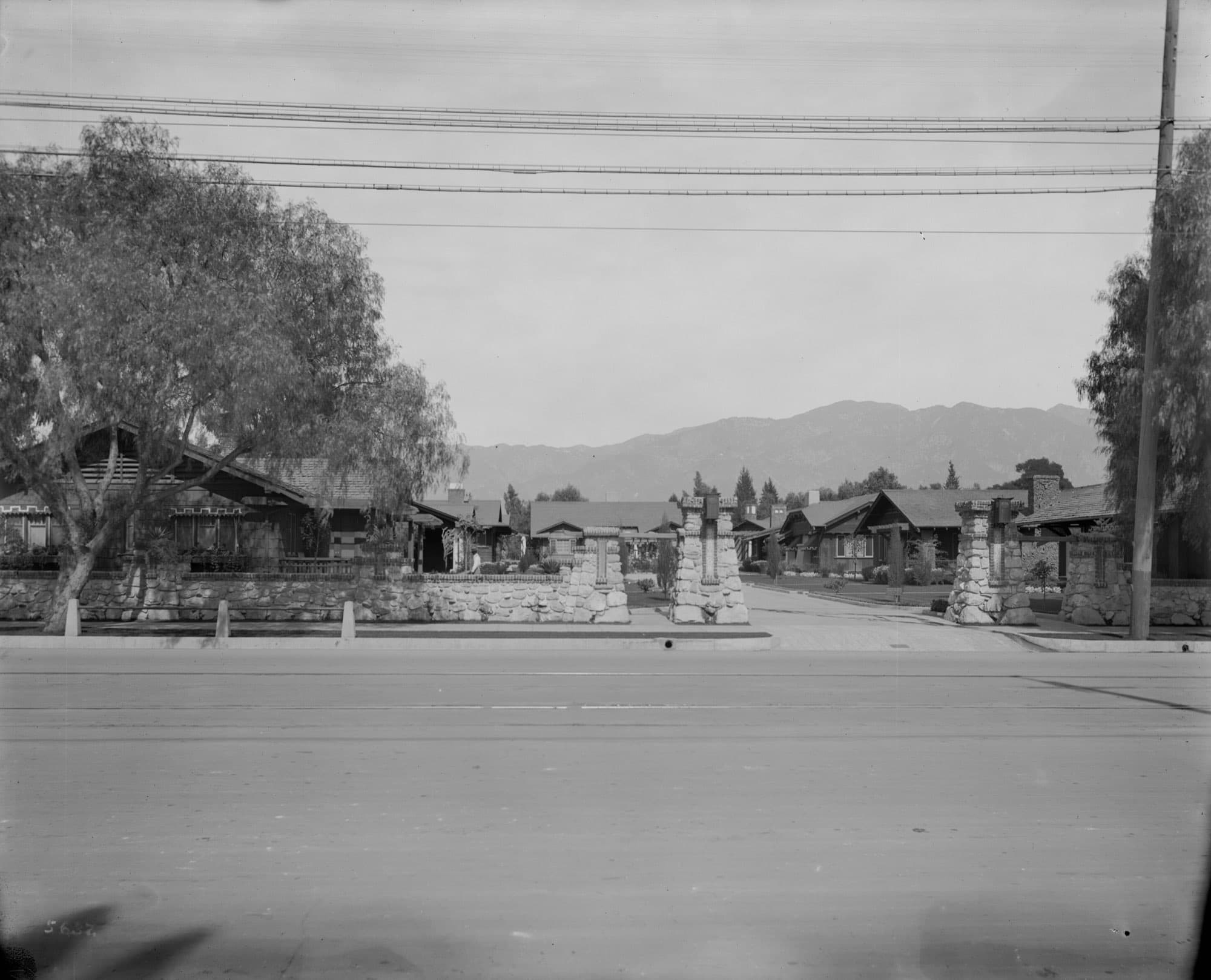 Black-and-white view across a street of stone gate piers and a driveway into a court of low shingled bungalows, with mountains behind