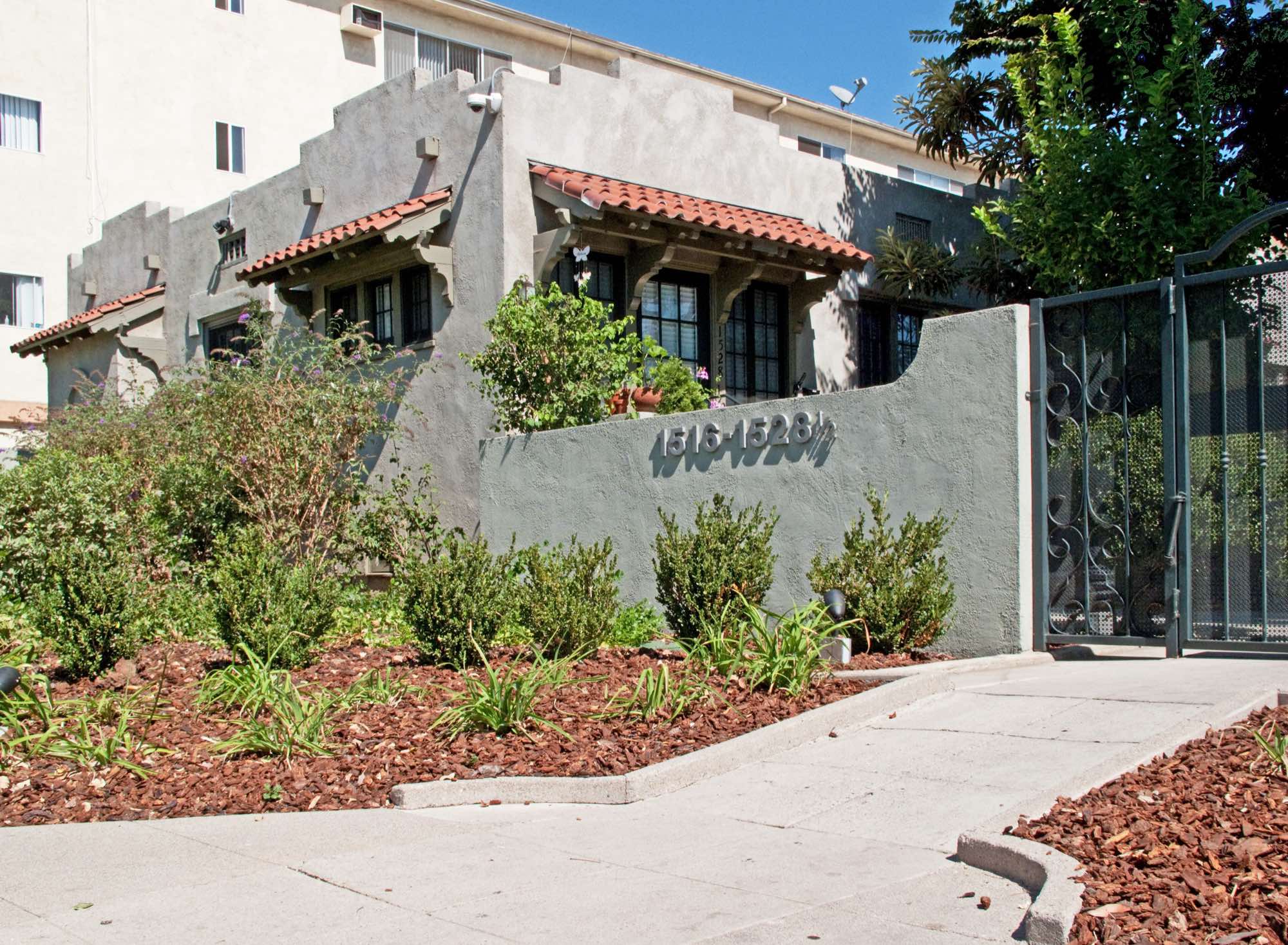 Grey stucco bungalow court with red-tile window hoods behind a low wall, an apartment block rising behind it in East Hollywood