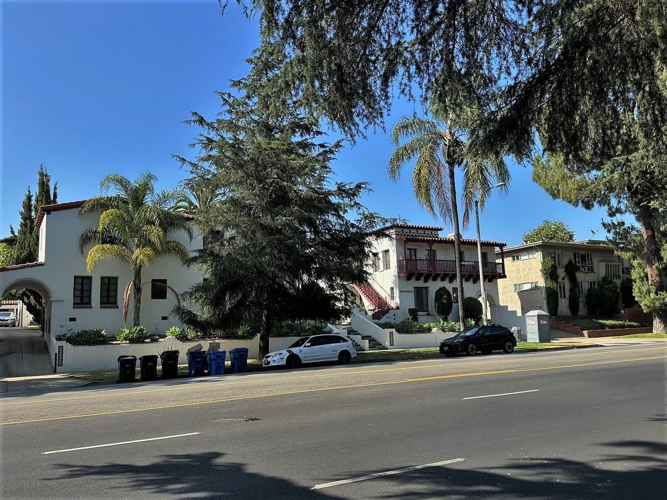 Two-storey white Spanish Revival courtyard apartments with red-tile roofs and a wooden balcony behind palms on Los Feliz Boulevard