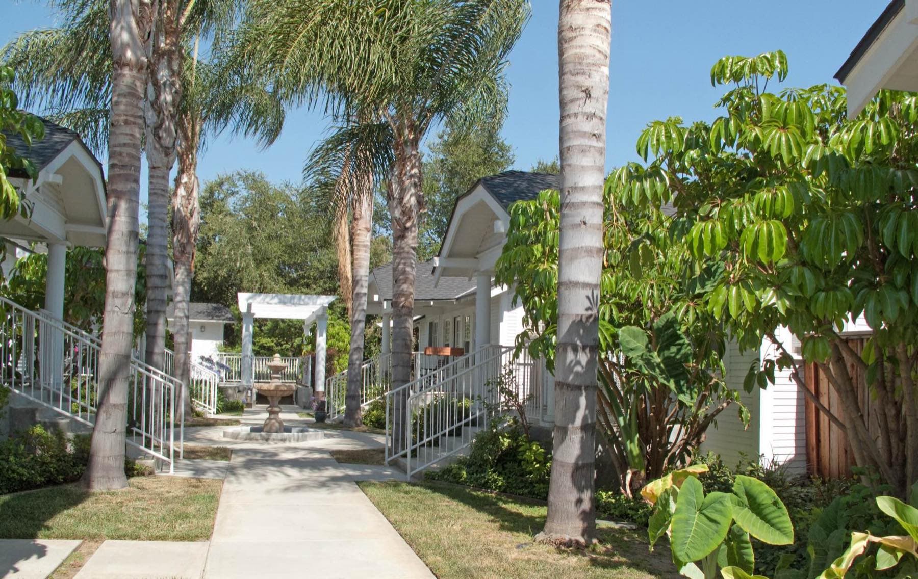 Bonnie Court in Pasadena, a classic of missing middle housing: a concrete path between two rows of white clapboard bungalows with gabled porches, under tall palms, leading to a fountain and pergola